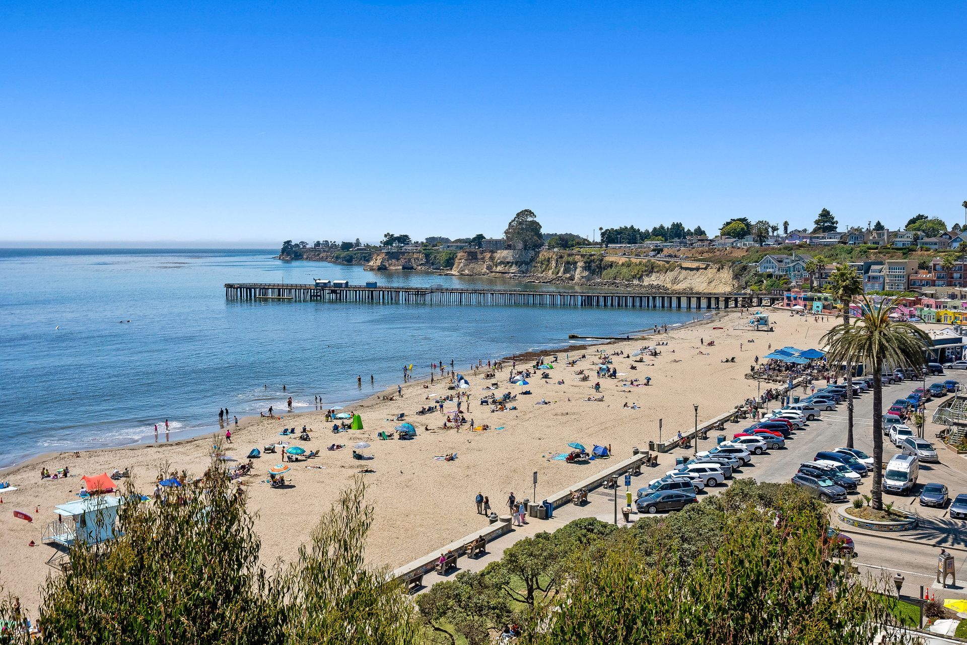 Sunny beach with people on the sand, ocean waves, and a pier under a clear blue sky