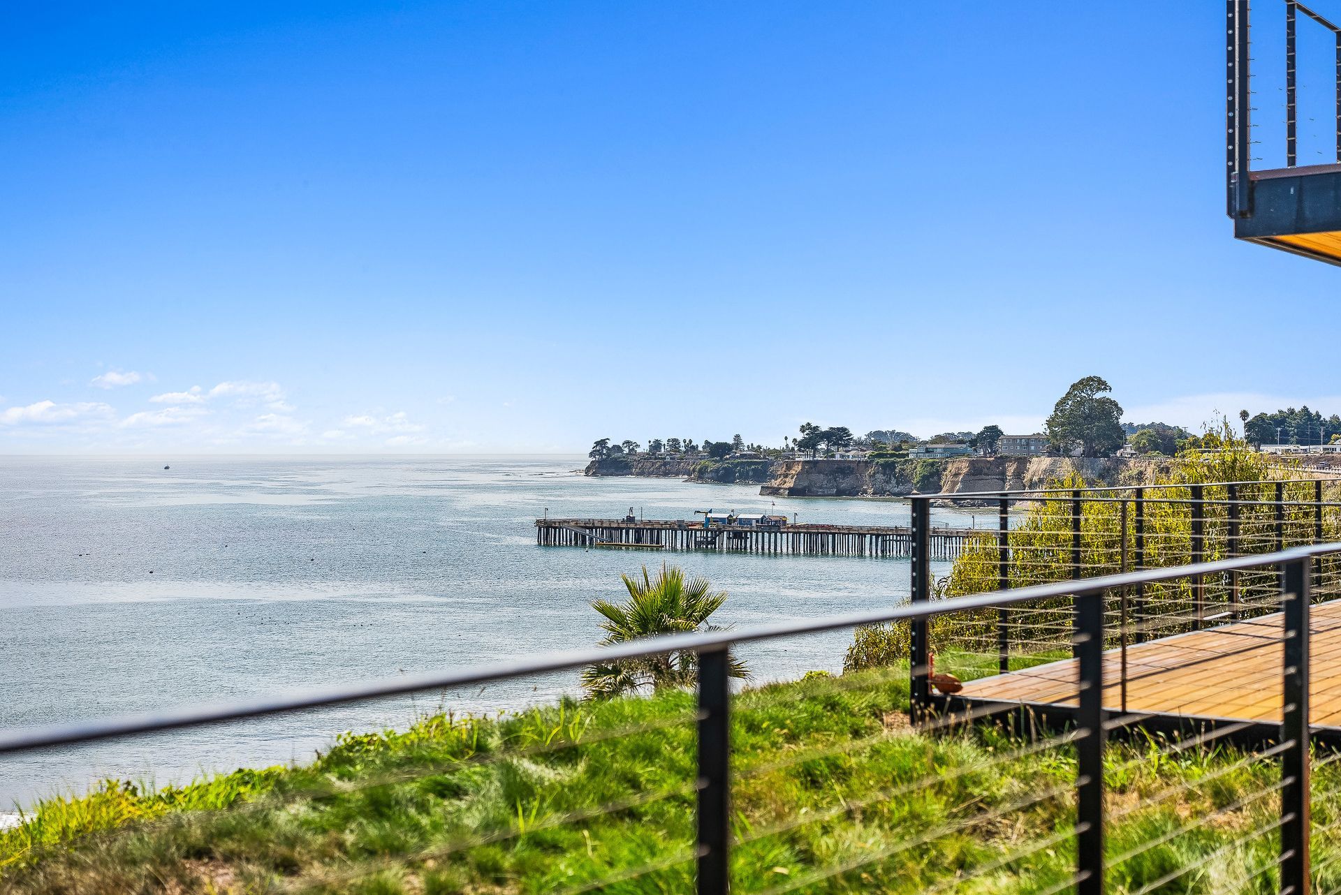 Sunny waterfront view with grassy shoreline, pier, and distant houses under a blue sky