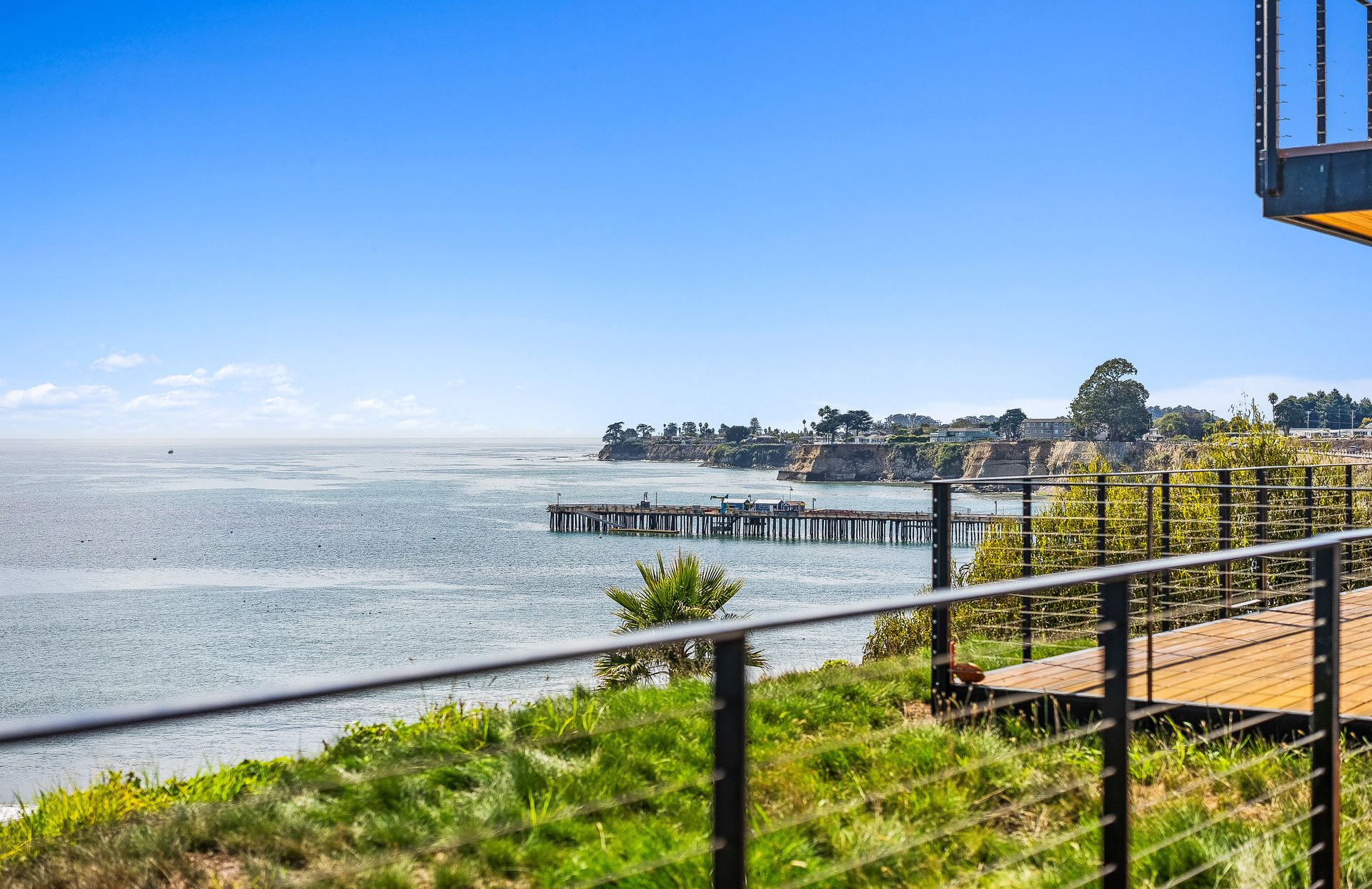 Sunny waterfront view with a pier, grassy shore, and black fence under a blue sky