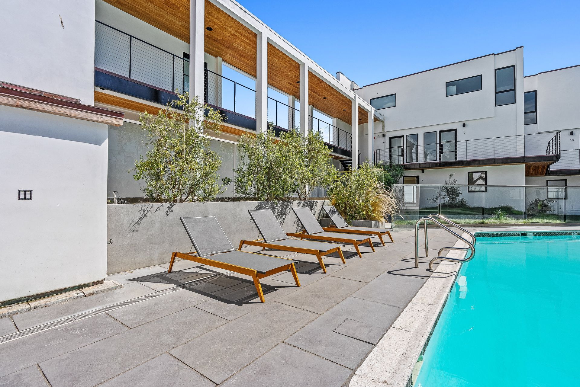 Modern poolside lounge area with wooden chairs and a turquoise swimming pool beside white buildings