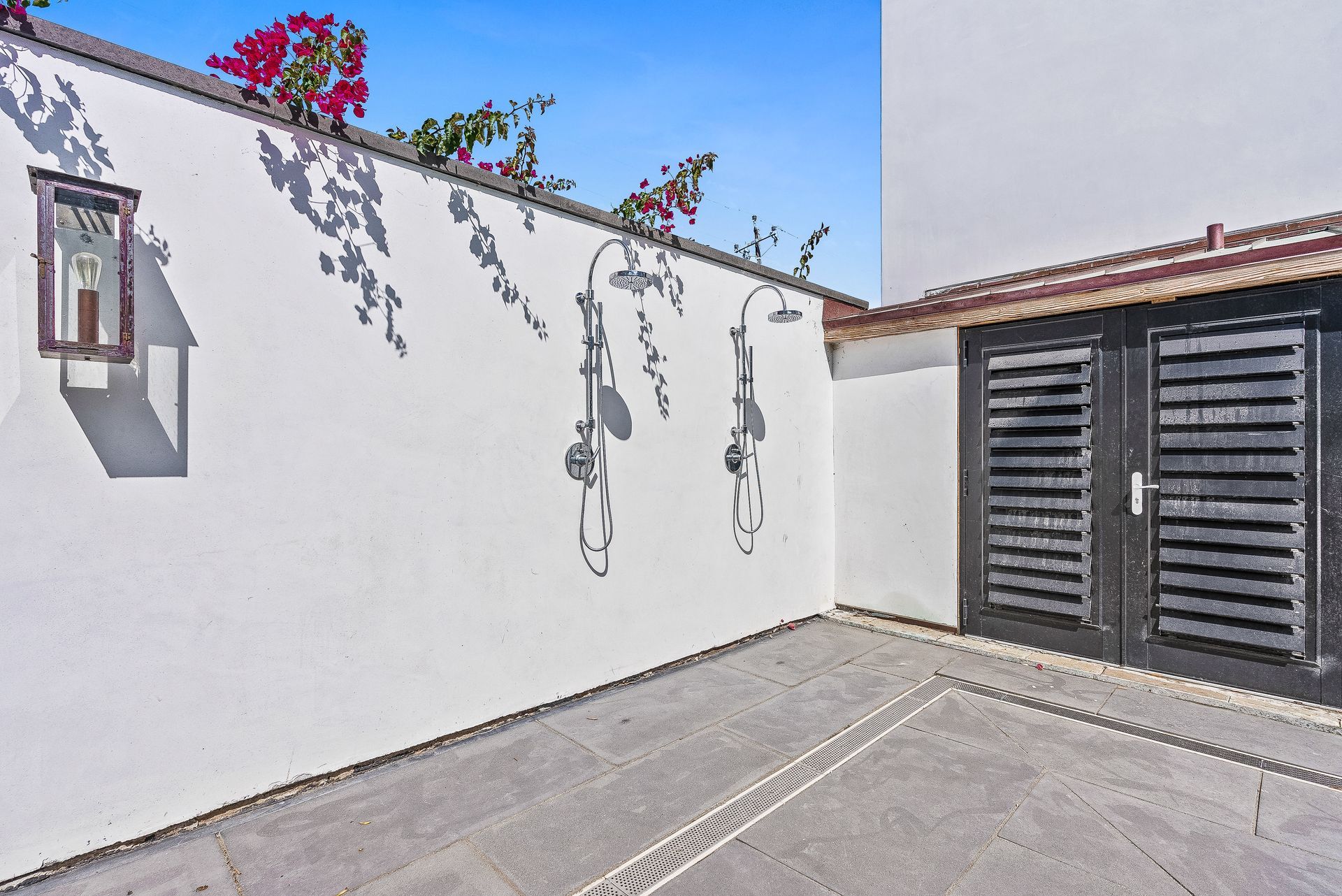 Sunlit courtyard with white walls, gray paving, and a dark wooden gate beside flowering vines.