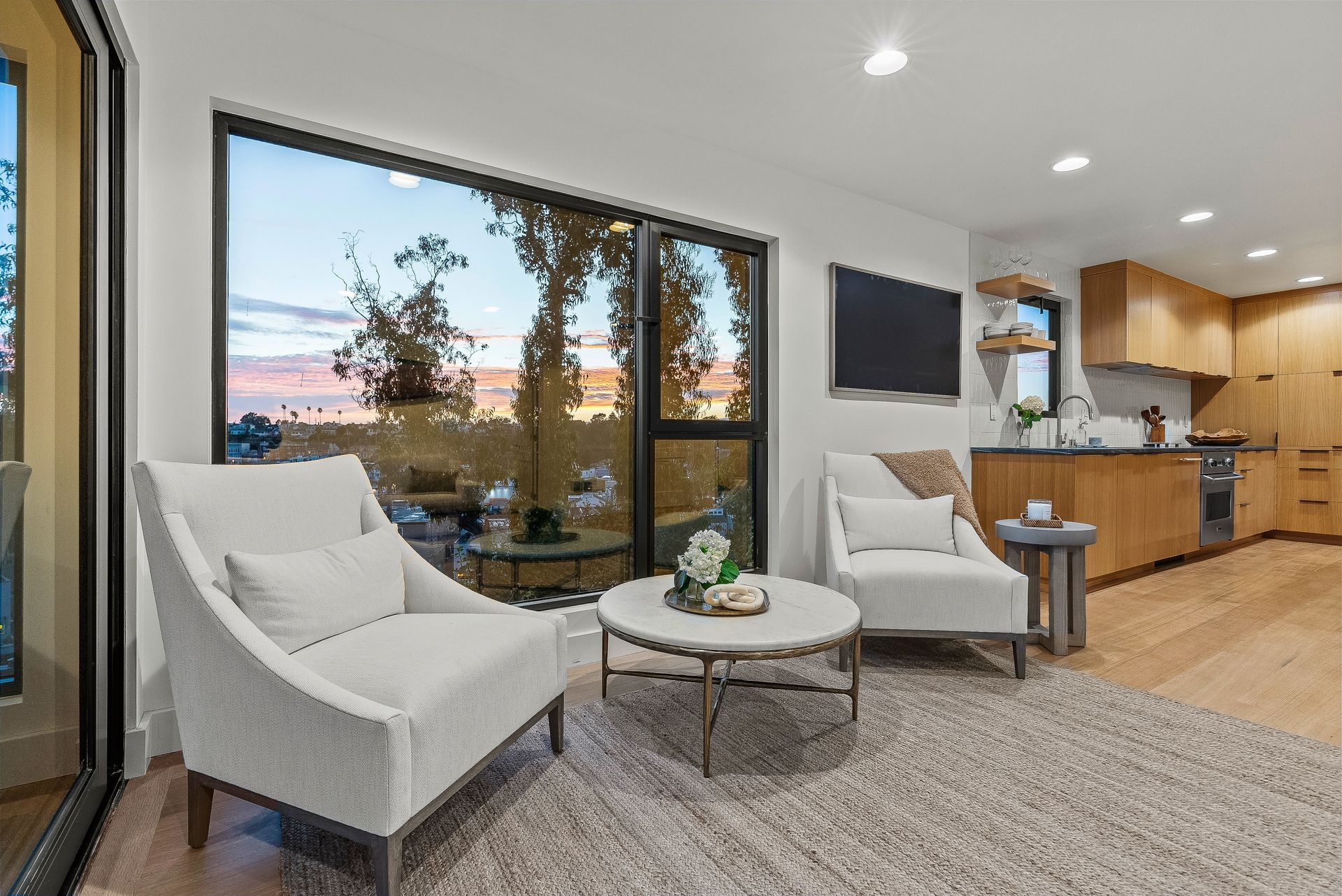 Modern living room with two white chairs, round table, large window, and open kitchen view