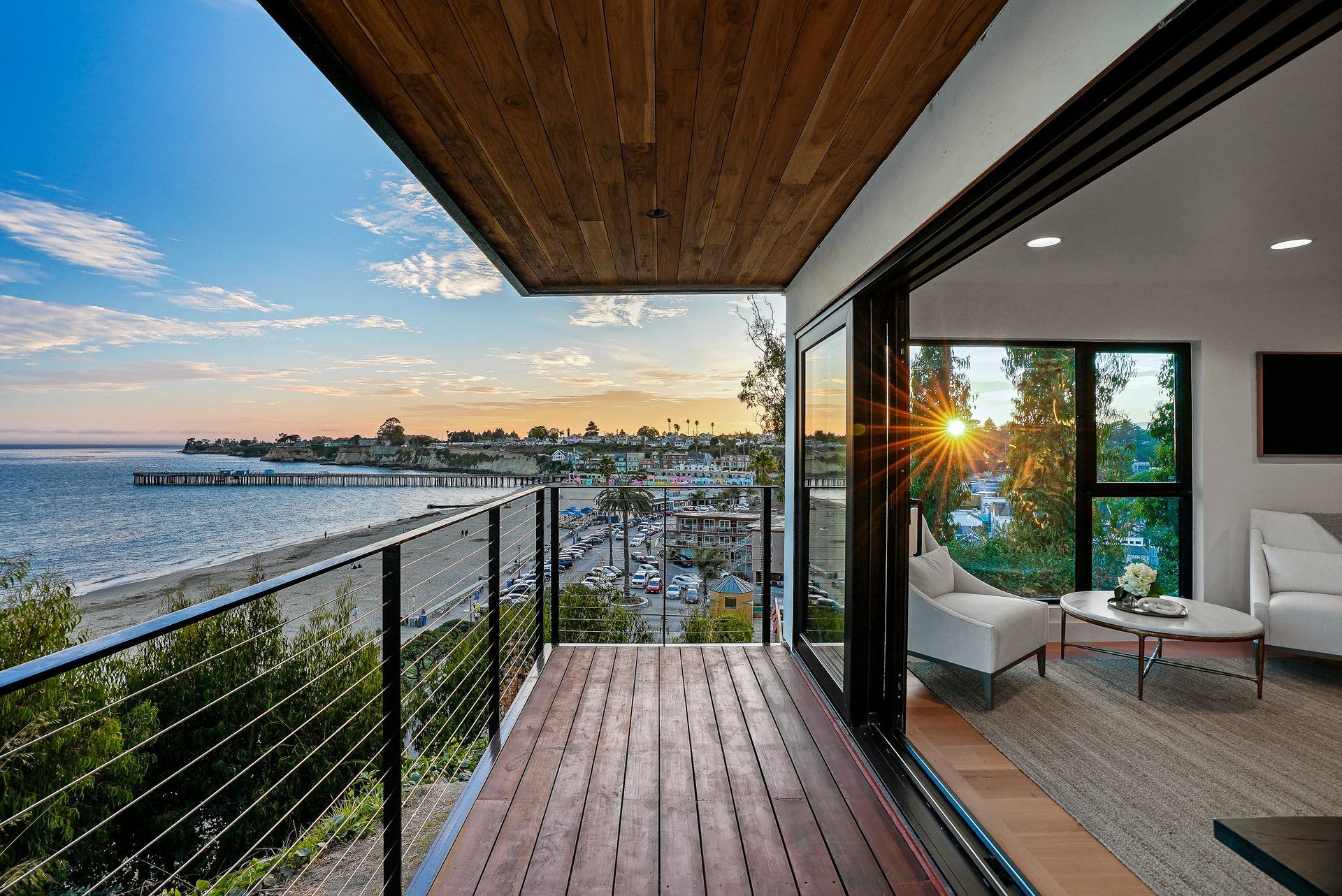 Sunset ocean-view balcony with wooden deck, glass railing, and a modern indoor lounge beside it