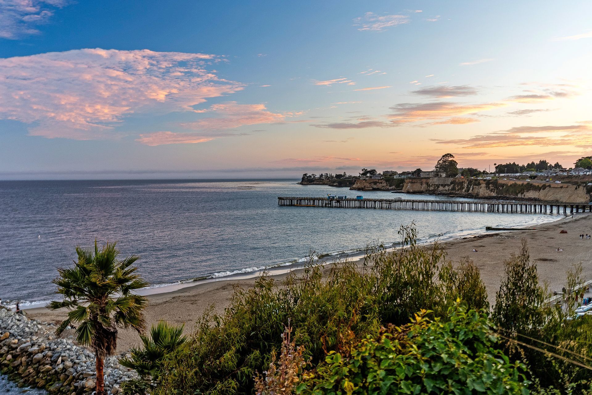 Coastal beach at sunset with calm ocean, pier, and palm trees under a pink-blue sky