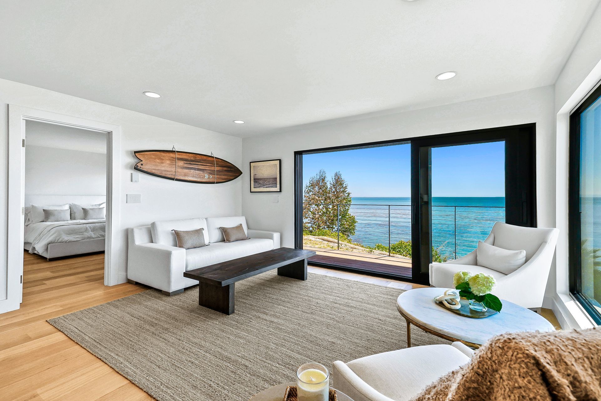 Modern living room with white furniture, ocean view through large windows, and surfboard wall decor