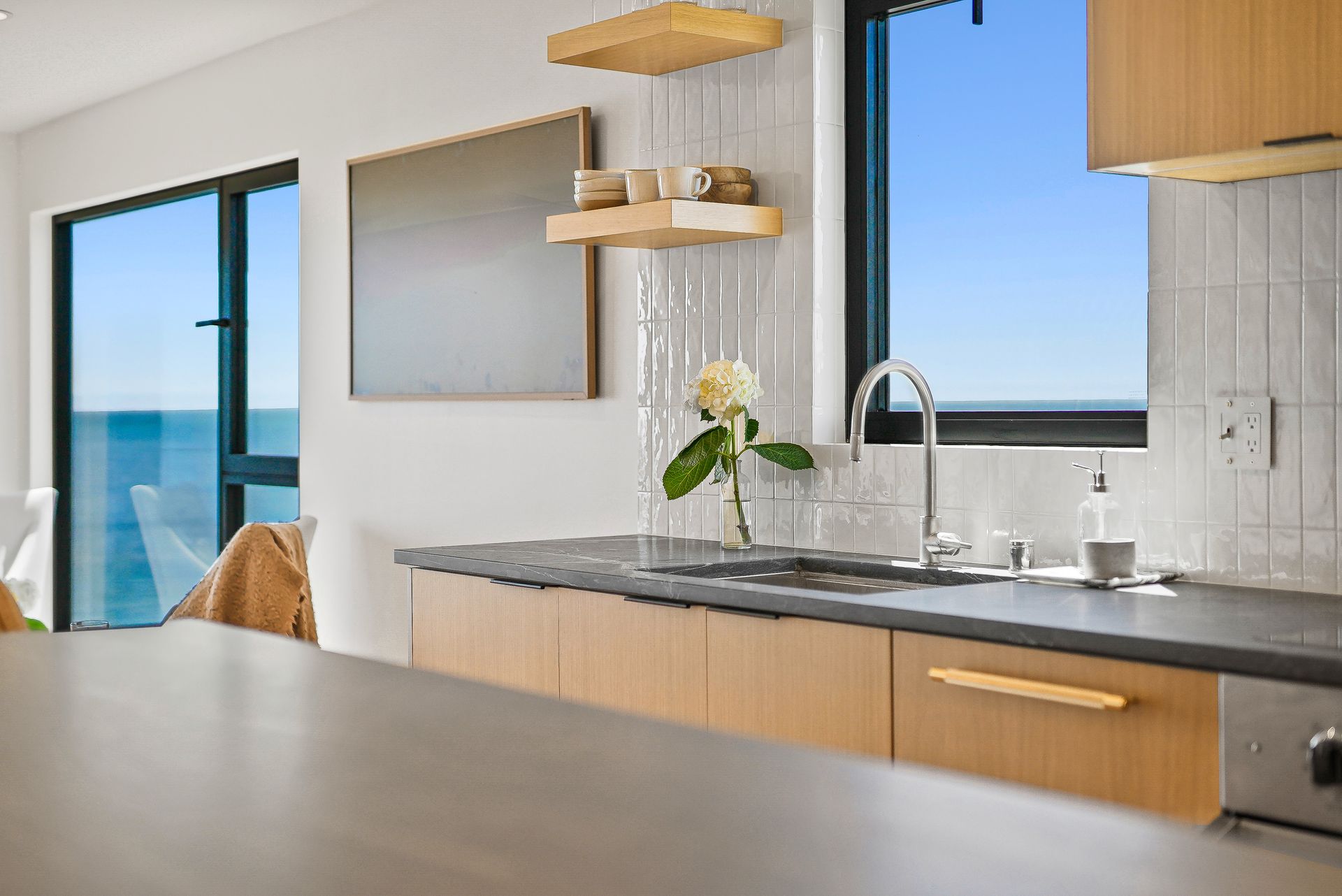 Modern kitchen with light wood cabinets, black countertops, and ocean view through large windows
