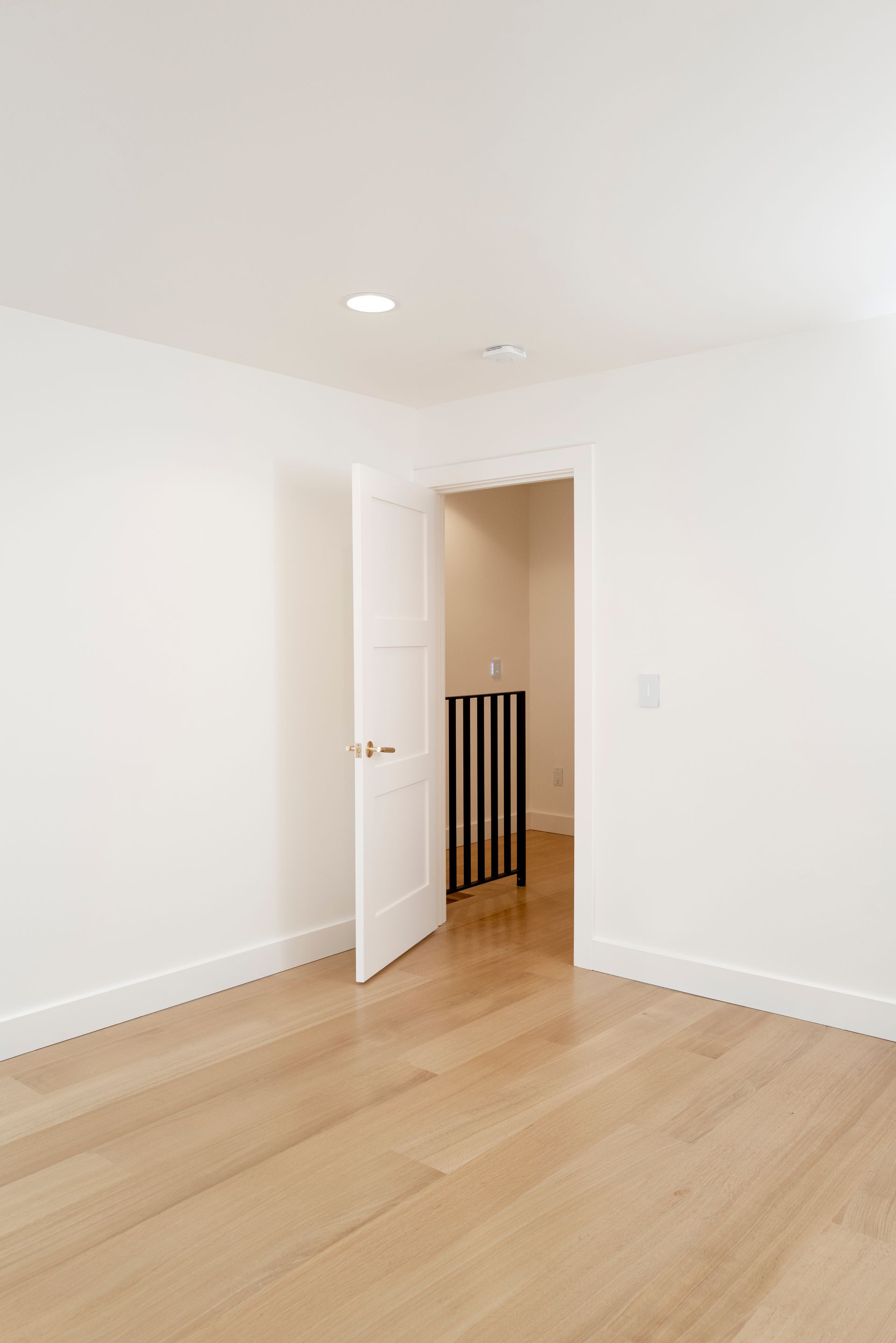 Empty white room with light wood floors and an open door leading to a hallway.