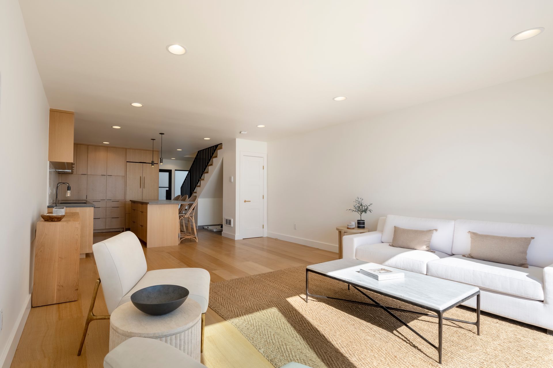 Bright open living room with white sofa, coffee table, kitchen, and stairs in the background