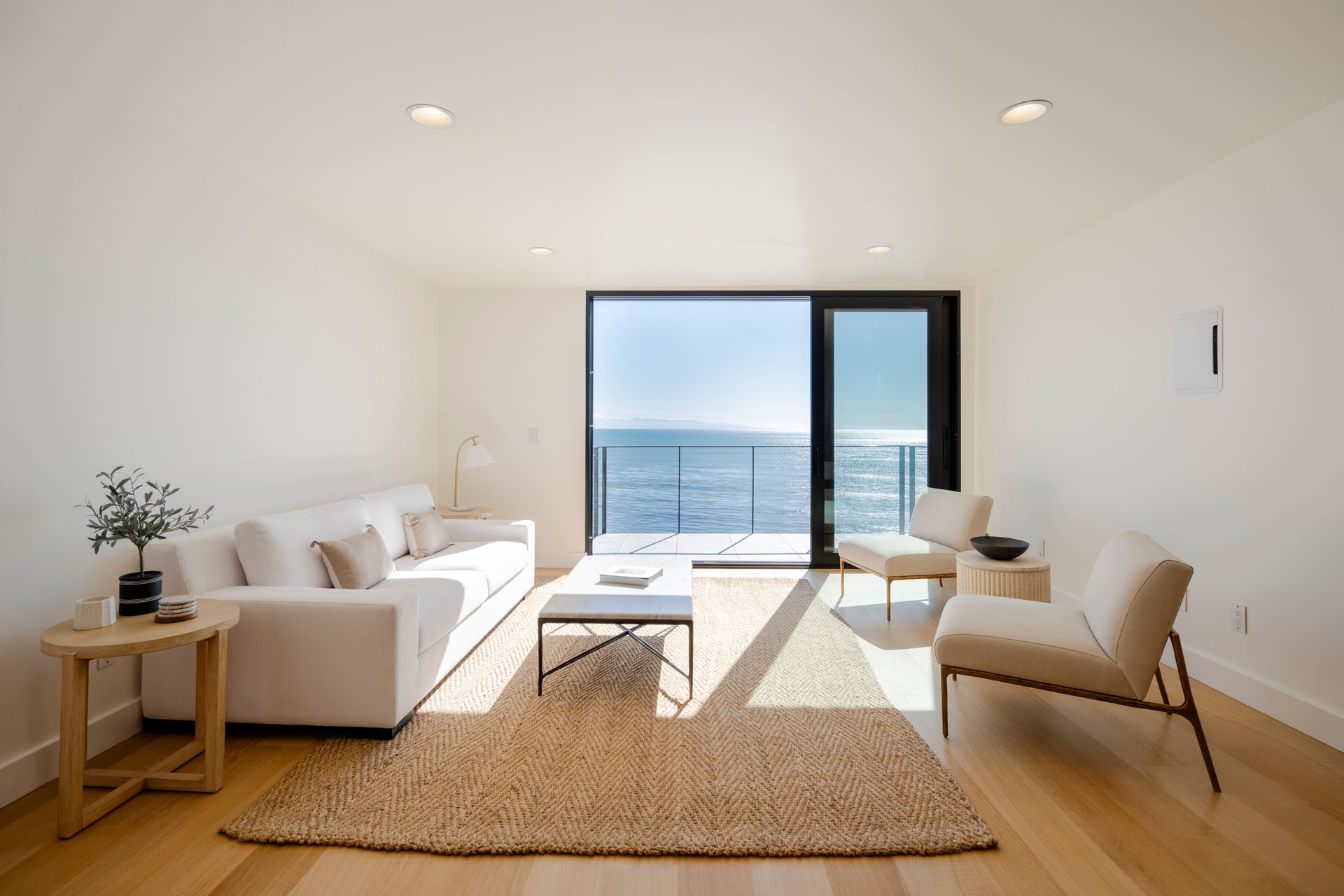 Bright minimalist living room with white furniture, wood floors, and ocean view through sliding glass doors