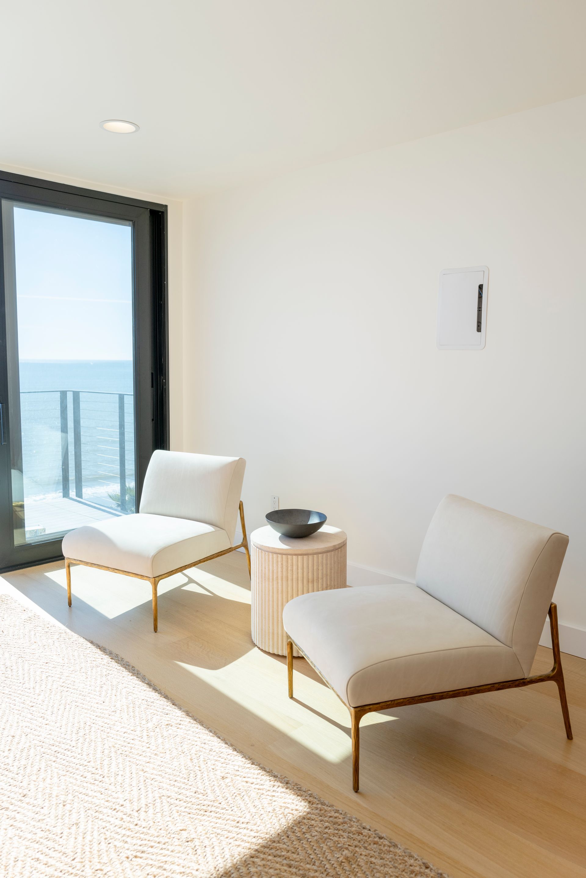 Bright minimalist room with two white lounge chairs, a small side table, and an ocean-view balcony door.