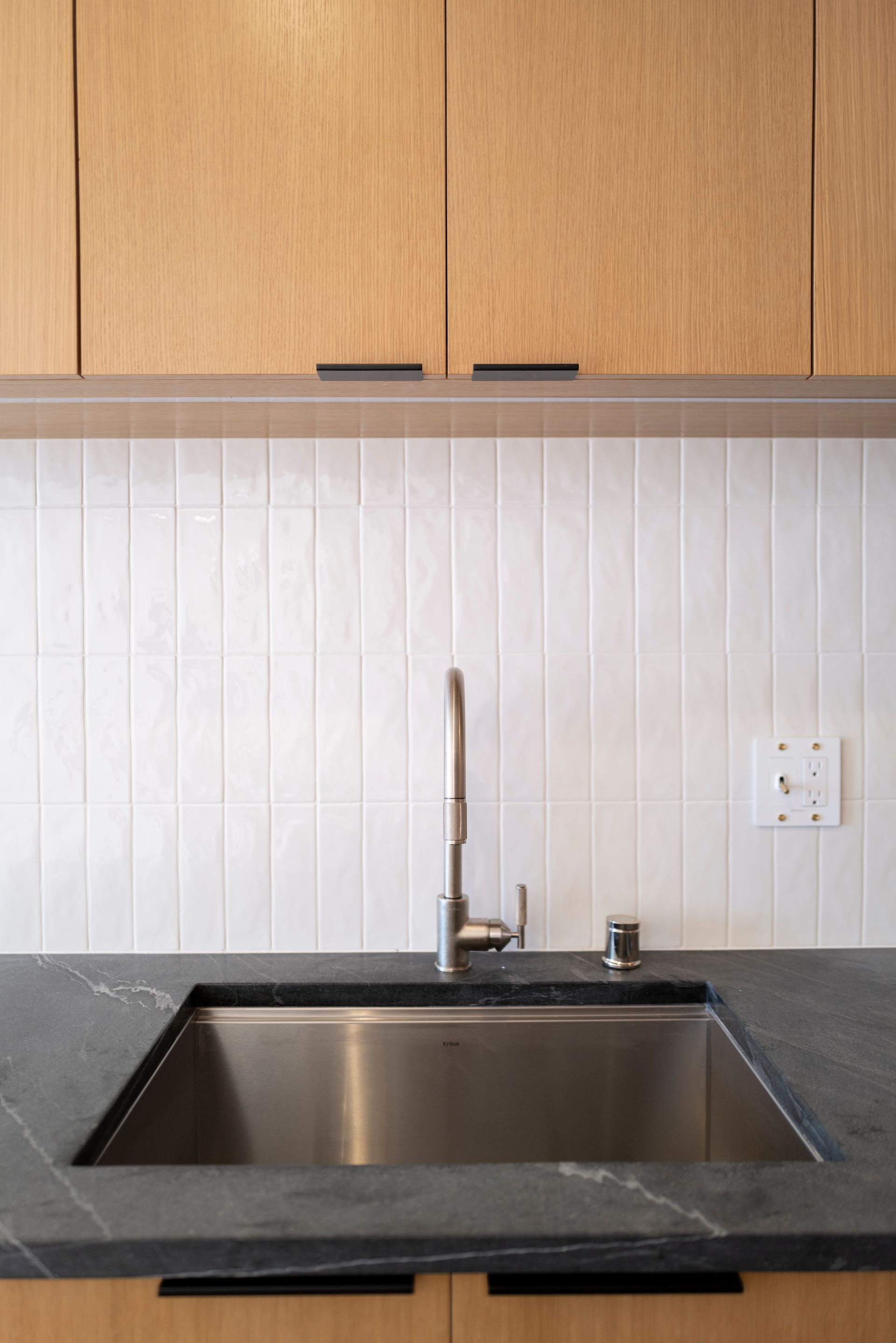 Modern kitchen sink with dark countertop, white backsplash, and light wood cabinets above