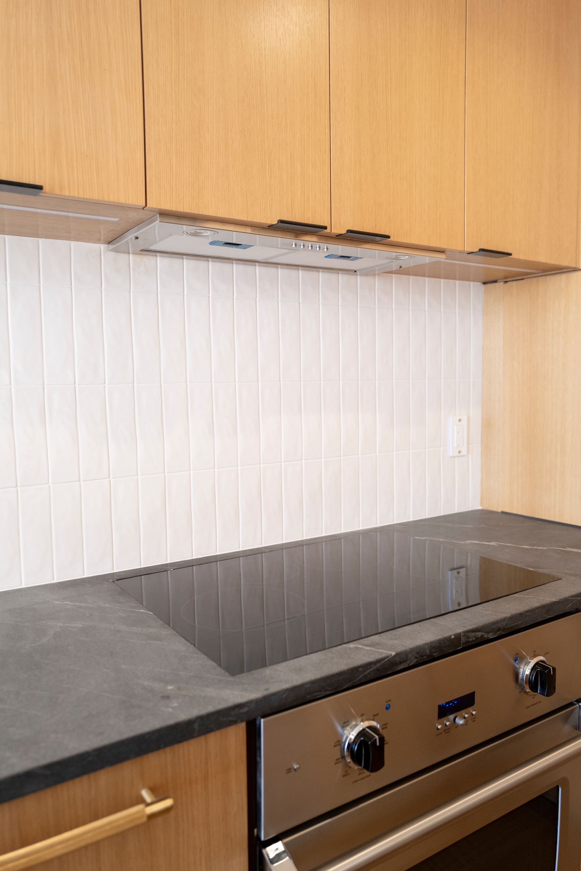 Modern kitchen with wood cabinets, white tiled backsplash, black countertop, and stainless steel oven.