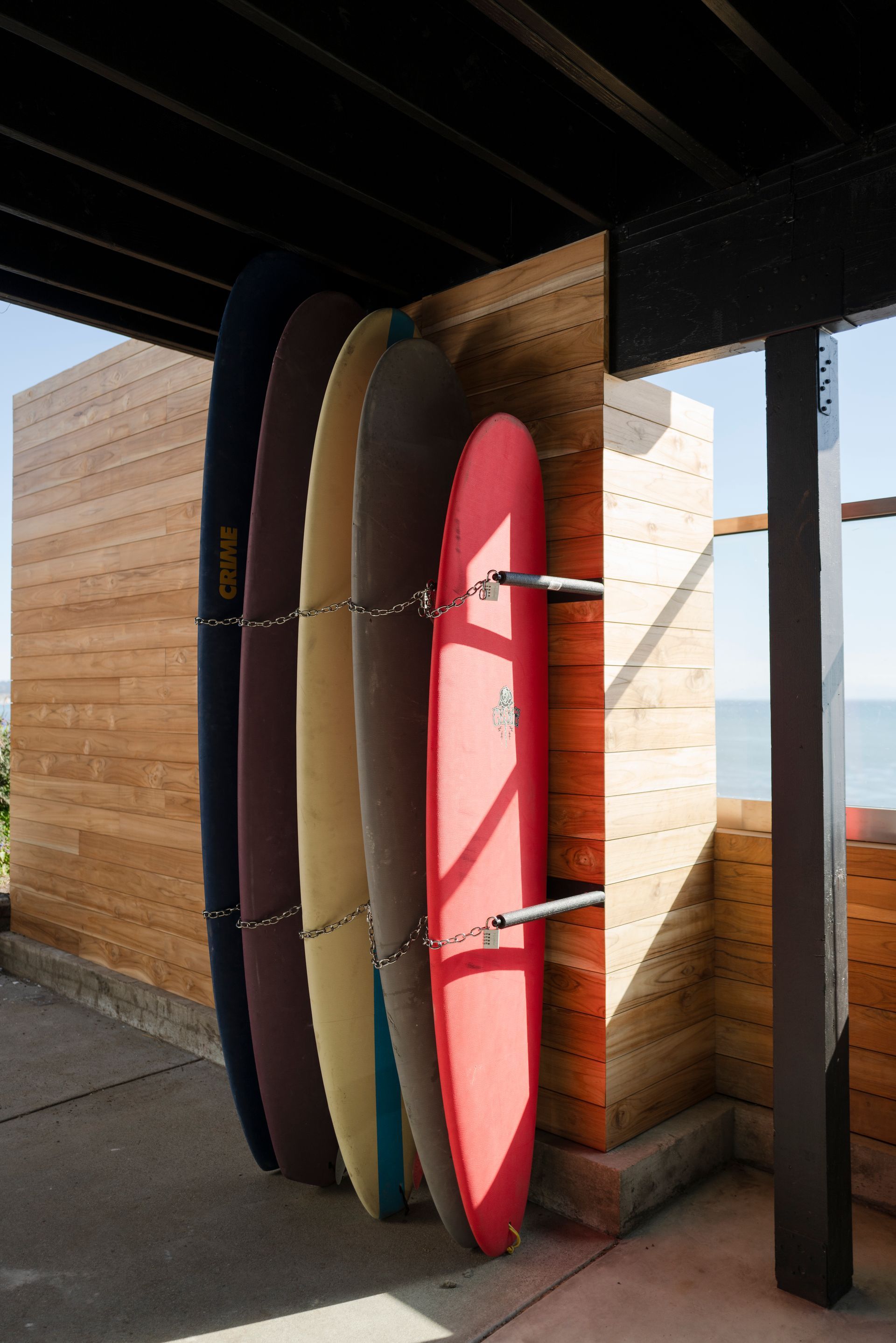 Surfboards stacked upright in a beachside rack beside a wooden wall and ocean view