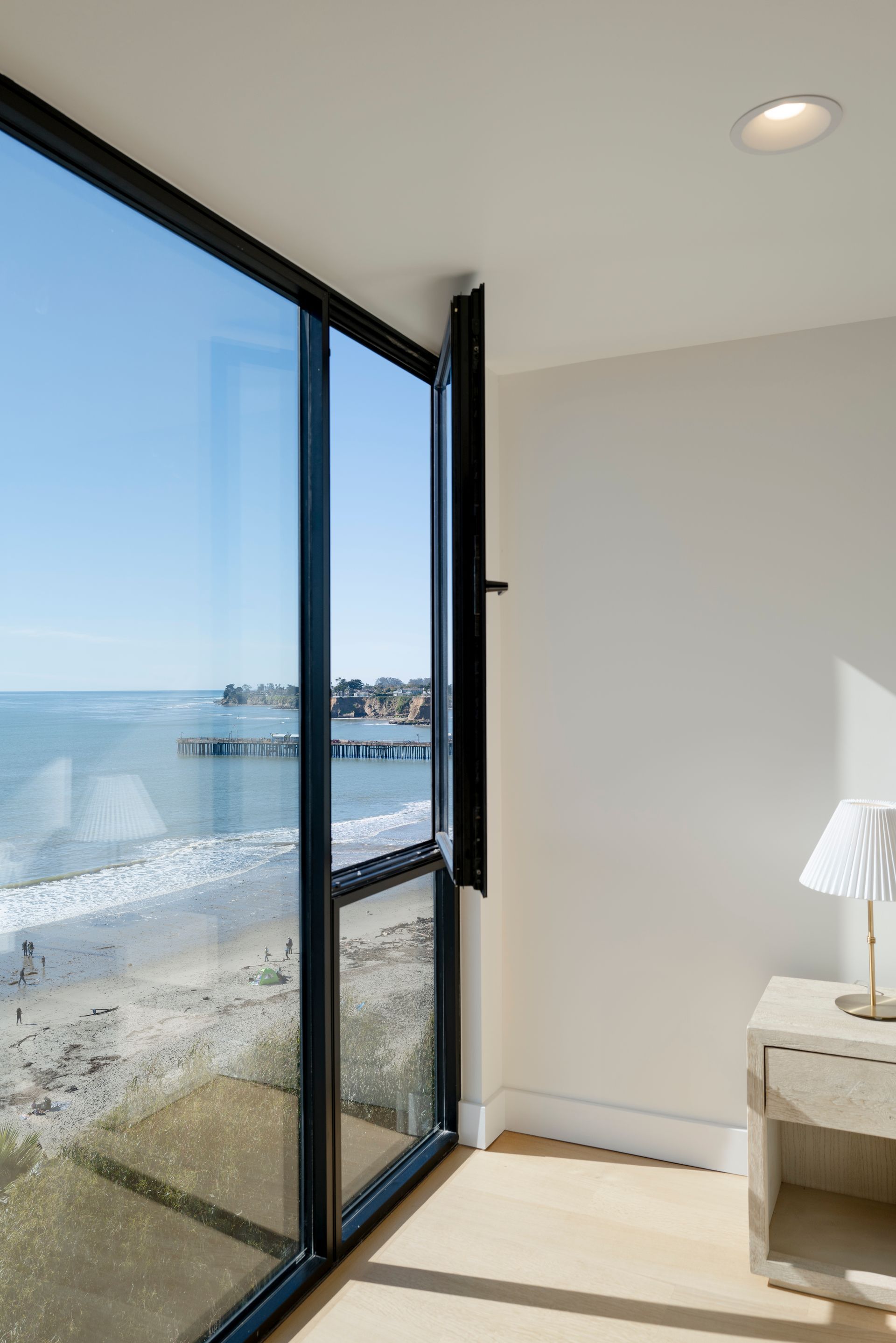 Sunlit room with floor-to-ceiling windows overlooking a beach and ocean, beside a small white table with lamp.
