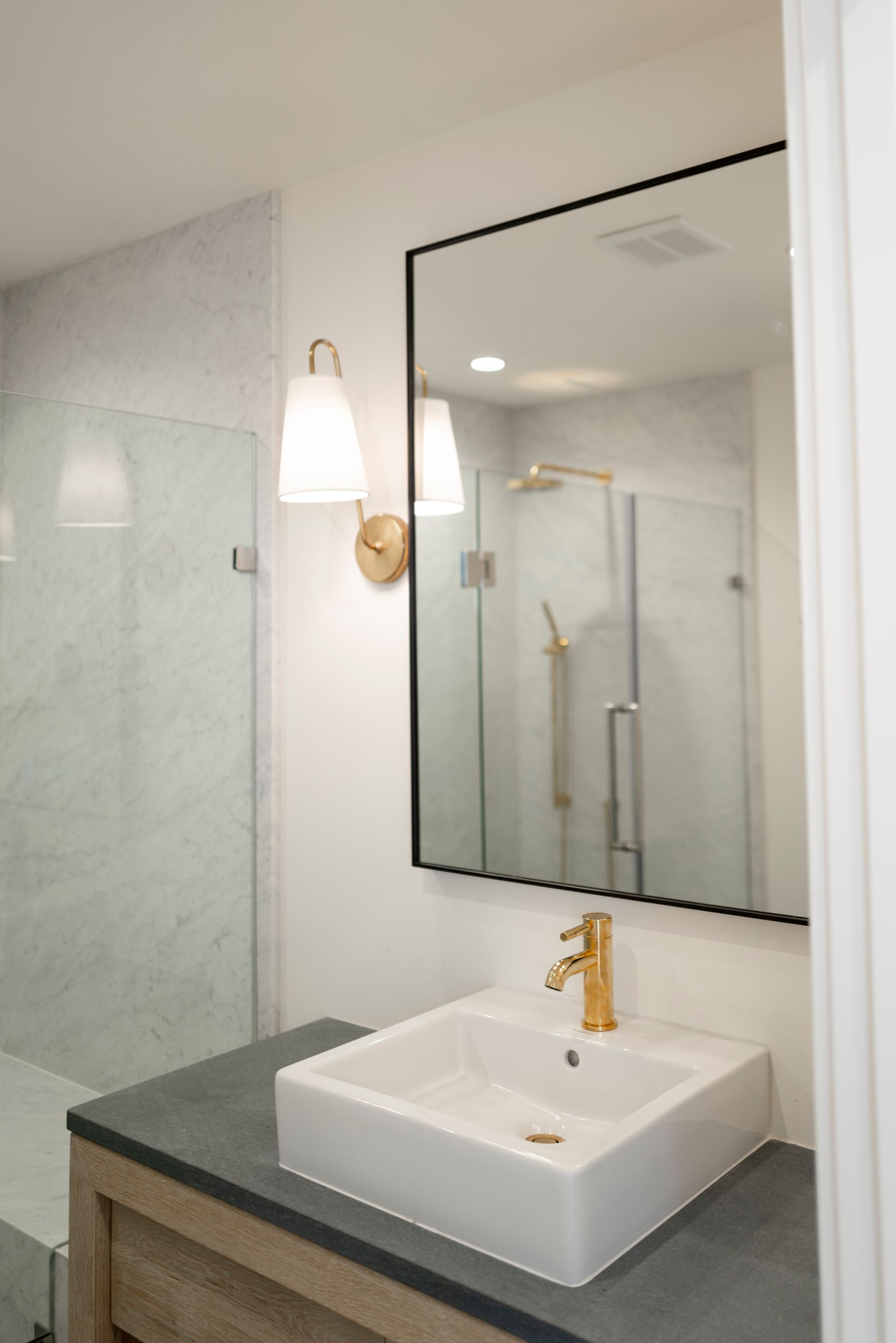 Modern bathroom vanity with white sink, gold faucet, and large wall mirror beside a glass shower.
