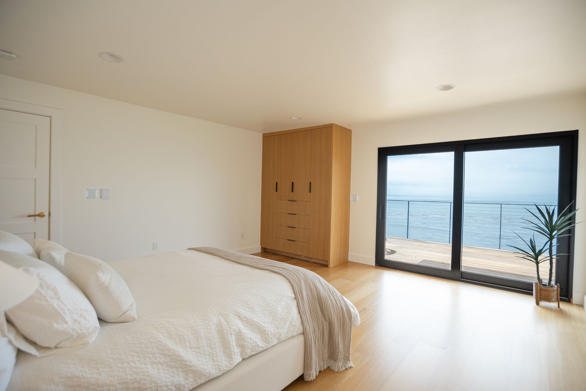 Bright bedroom with white bed, wood floor, wardrobe, and ocean-view sliding doors.