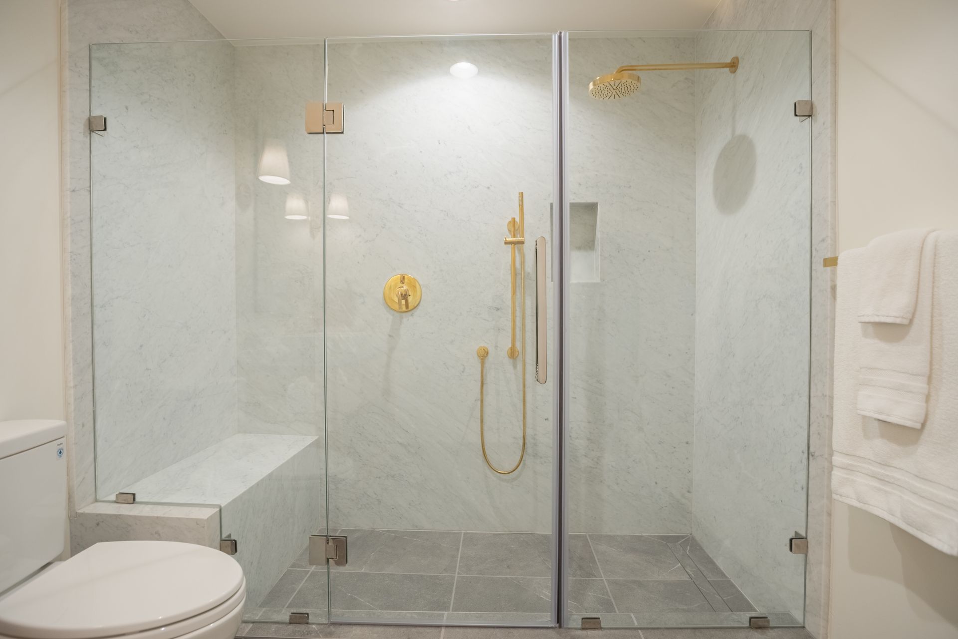 Accessible bathroom with glass shower, bench, gold fixtures, and towels on the wall