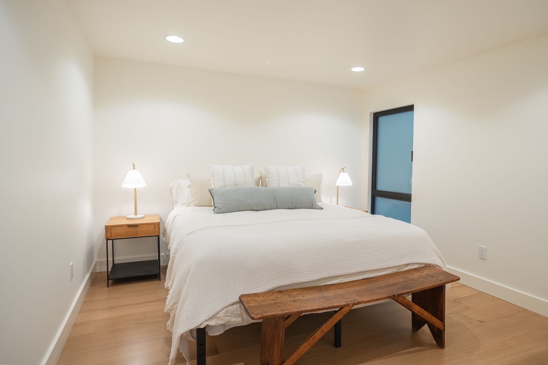 Bright bedroom with white bed linens, wooden bench, bedside lamps, and a frosted glass door
