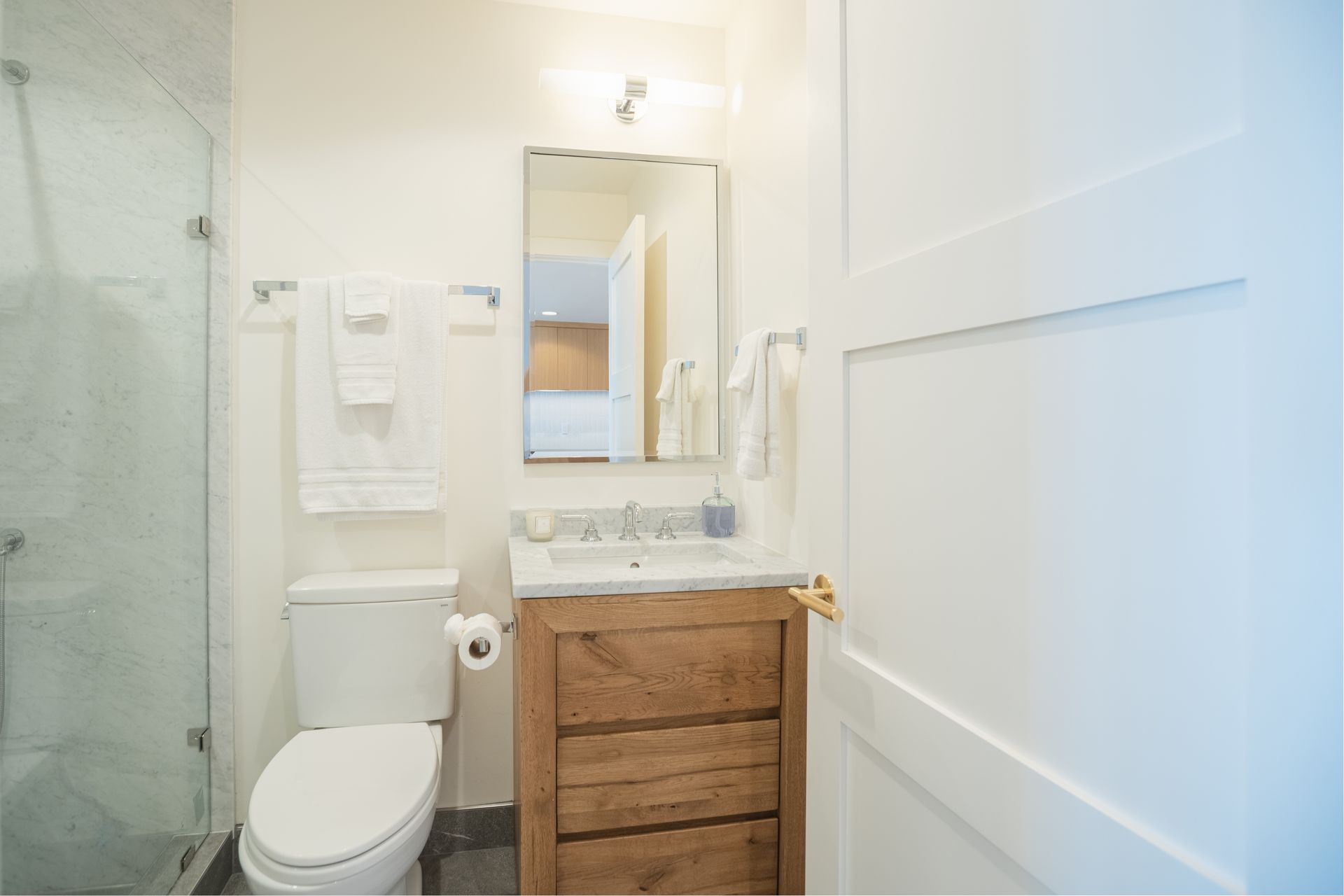 Small white bathroom with toilet, wood vanity, mirror, and glass shower door
