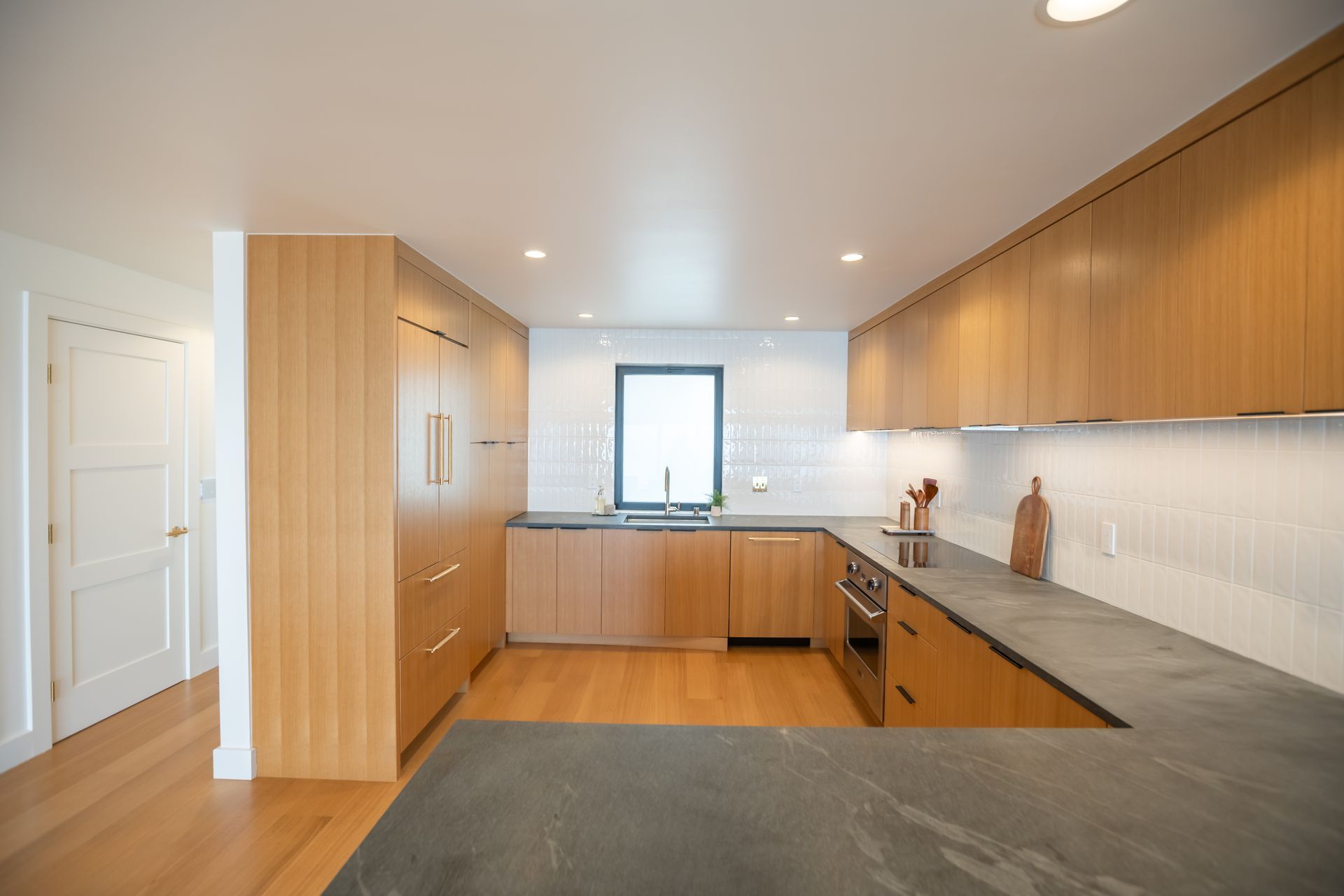 Modern kitchen with wood cabinets, gray countertops, white walls, and a window above the sink.