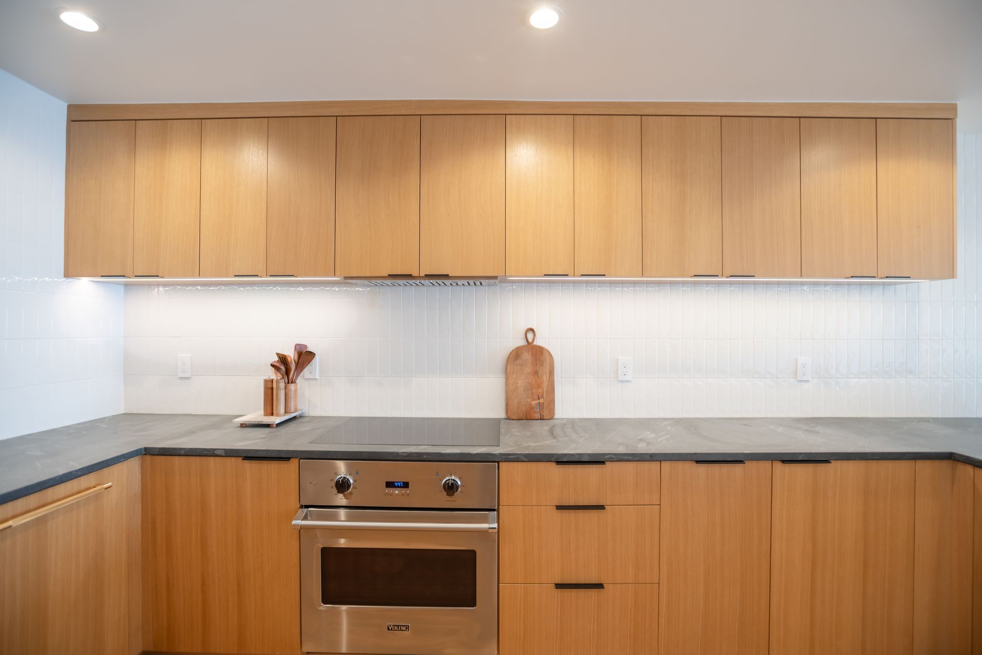 Modern kitchen with light wood cabinets, gray countertops, stainless steel oven, and white backsplash.
