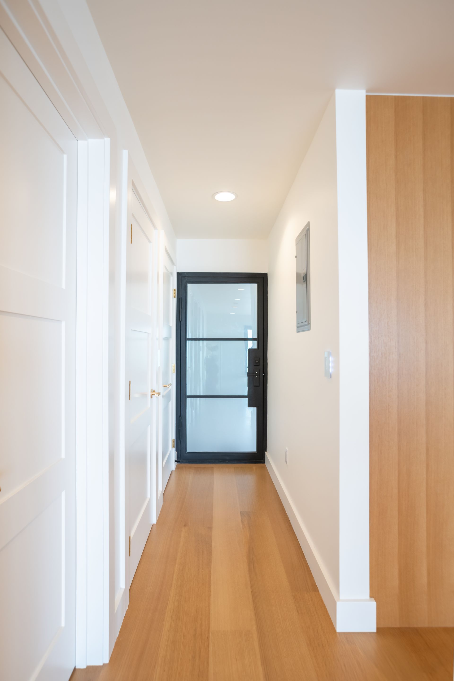 Narrow hallway with light wood floors, white walls, built-in closet doors, and a black-framed glass door at the end