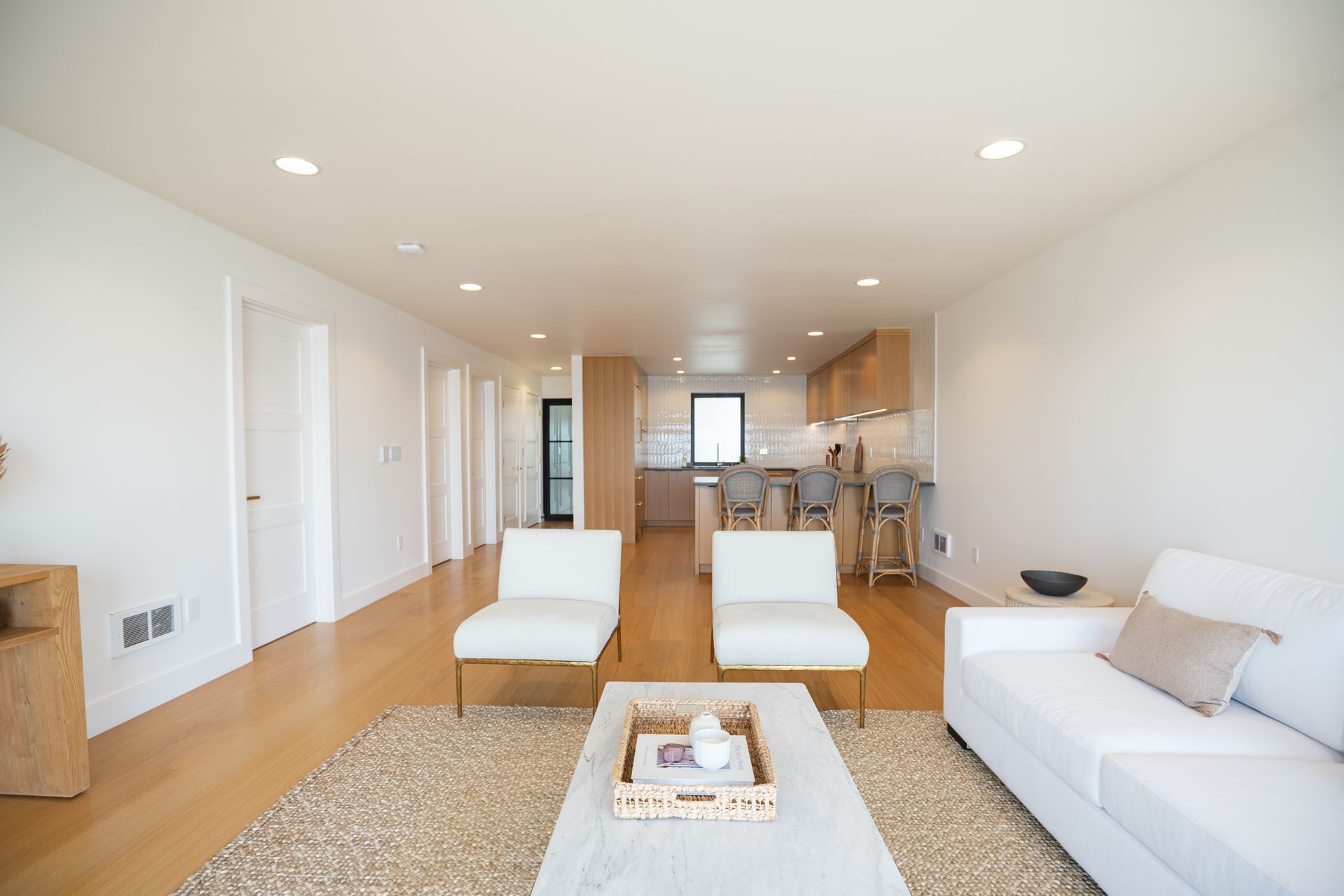 Bright open living room with white furniture, wood floors, rug, and kitchen area in the background