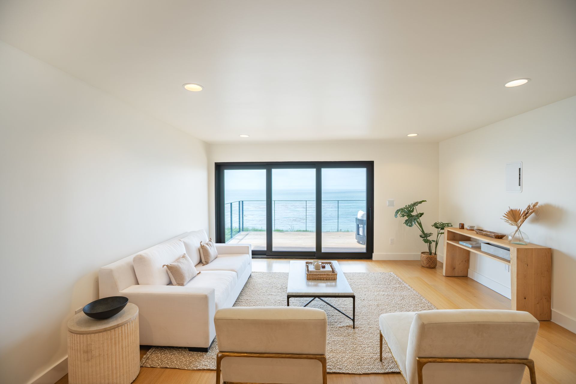 Bright modern living room with white sofa, beige chairs, large windows, and ocean view.