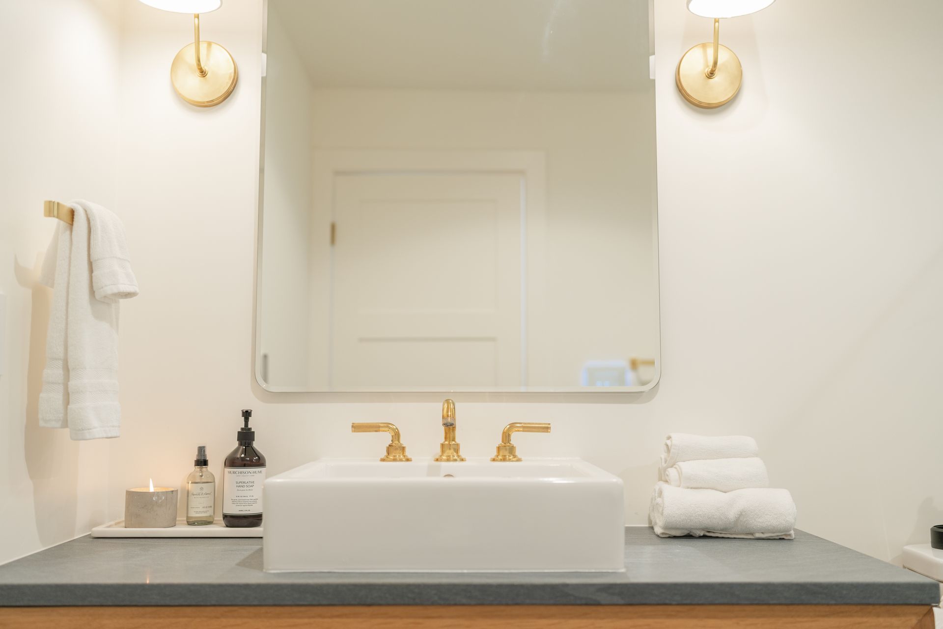 Modern bathroom vanity with white sink, gold faucet, mirror, and folded towels on a dark countertop