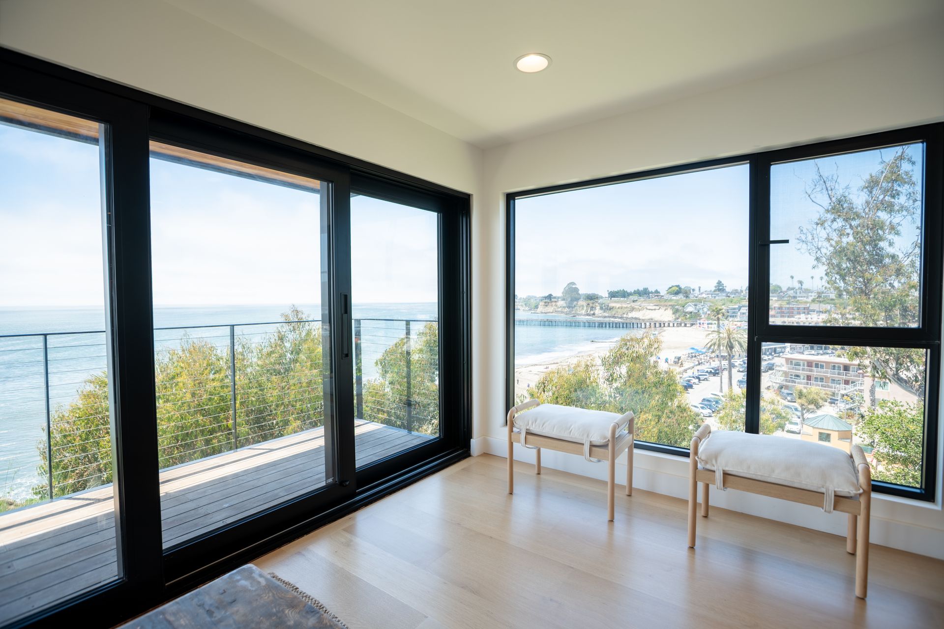 Bright living room with floor-to-ceiling windows overlooking water and a balcony, two white chairs by the view