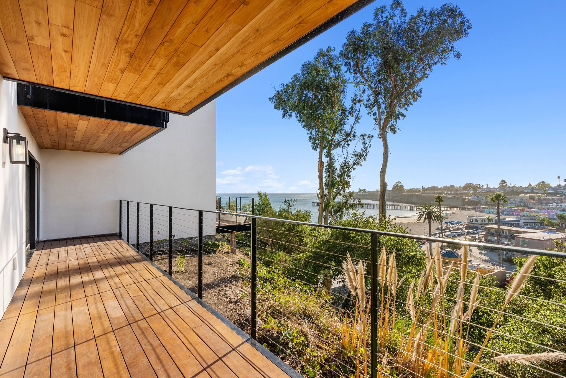 Modern wooden balcony with glass railing, white wall, trees, and a city view beyond the deck.