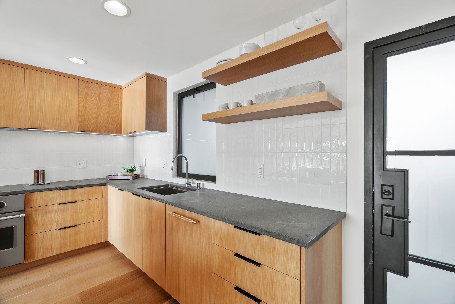 Modern kitchen with wood cabinets, dark counters, white tile backsplash, and floating shelves