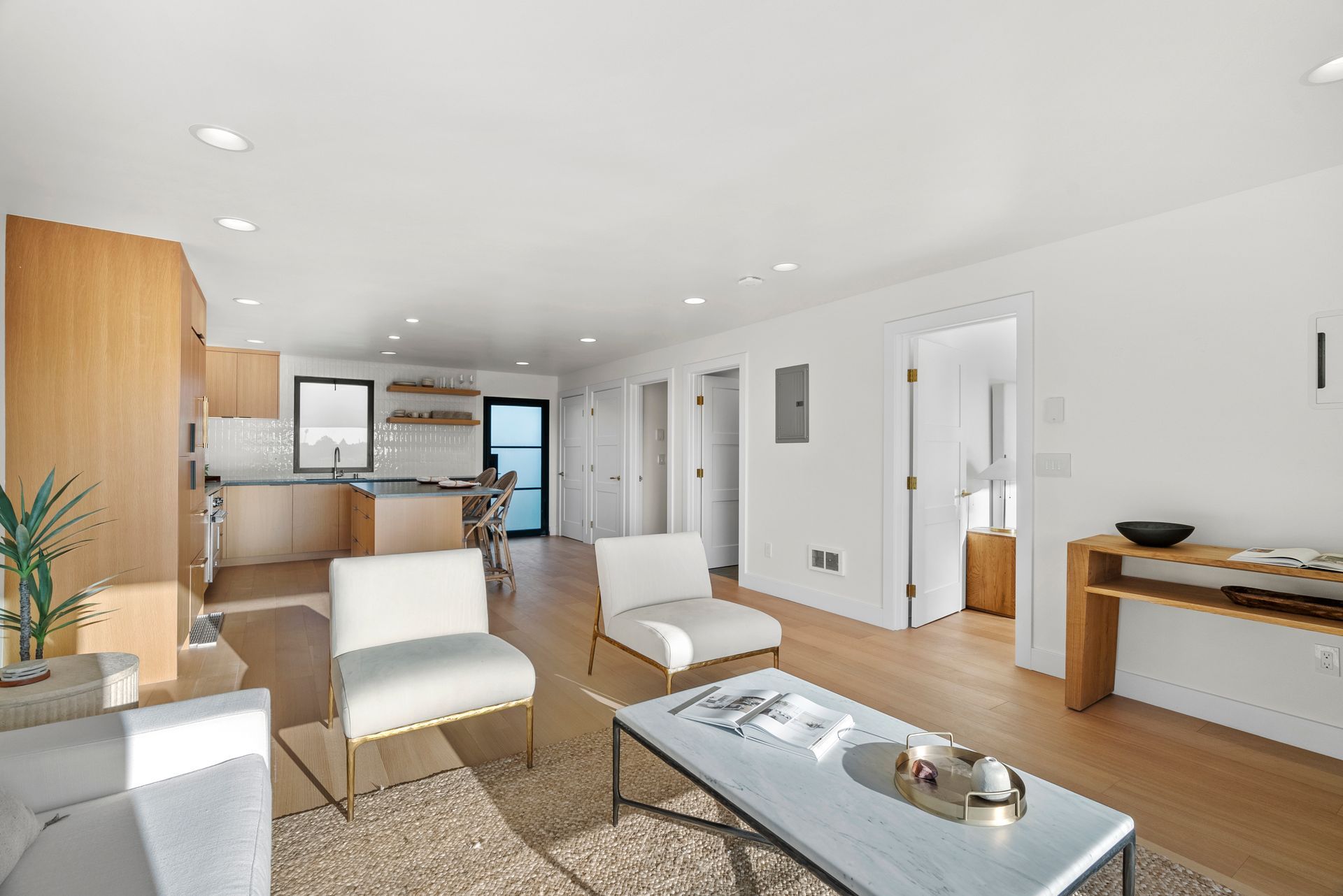 Bright, modern living room with white chairs, a coffee table, wood accents, and an open kitchen area.