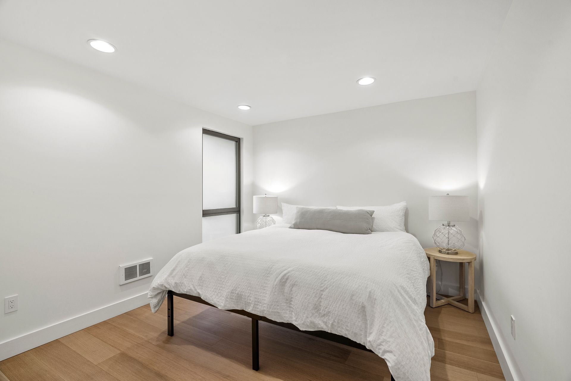 Minimal white bedroom with a bed, nightstands, lamp, and hardwood floor.