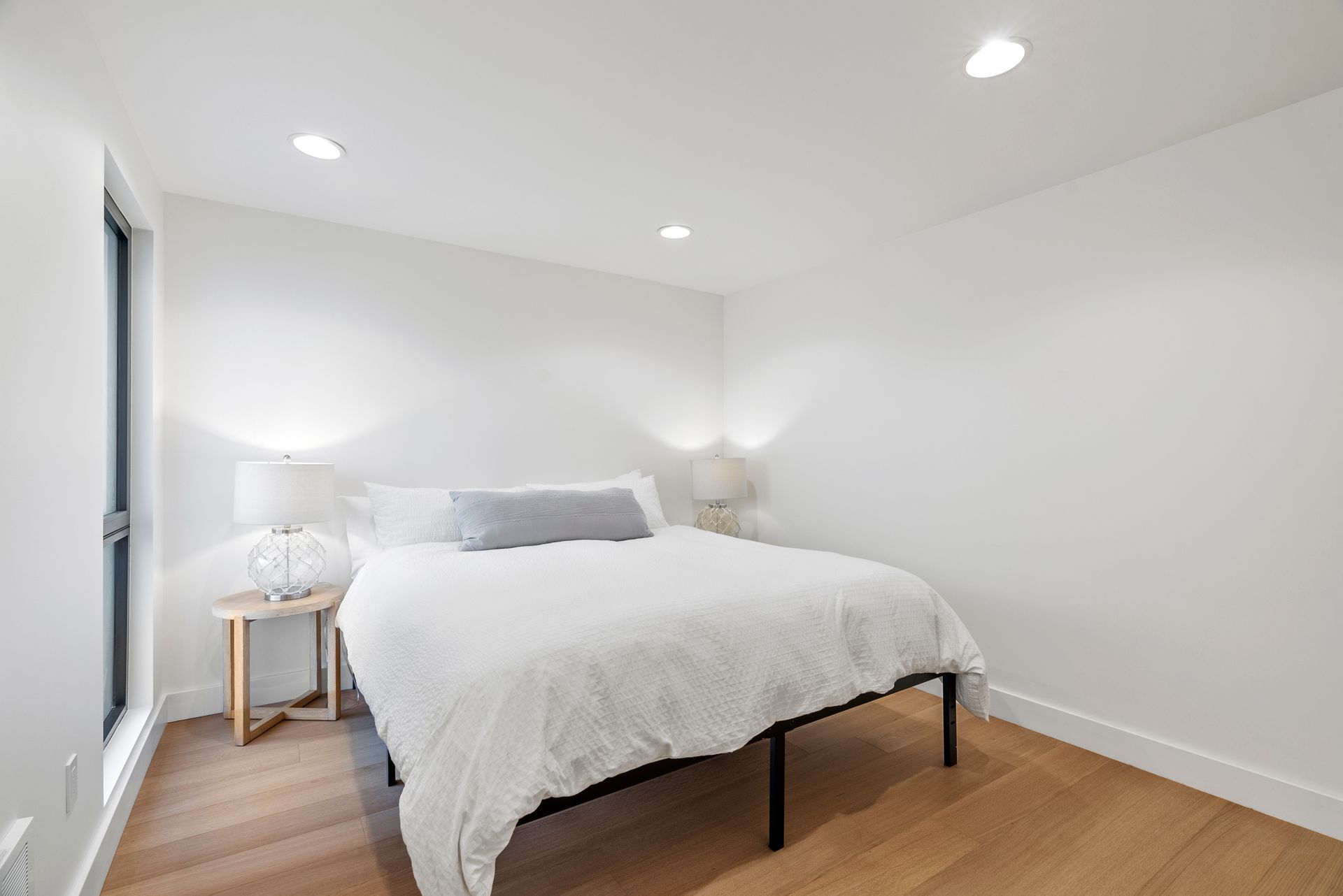 Bright white bedroom with a neatly made bed, wood floors, and recessed ceiling lights.