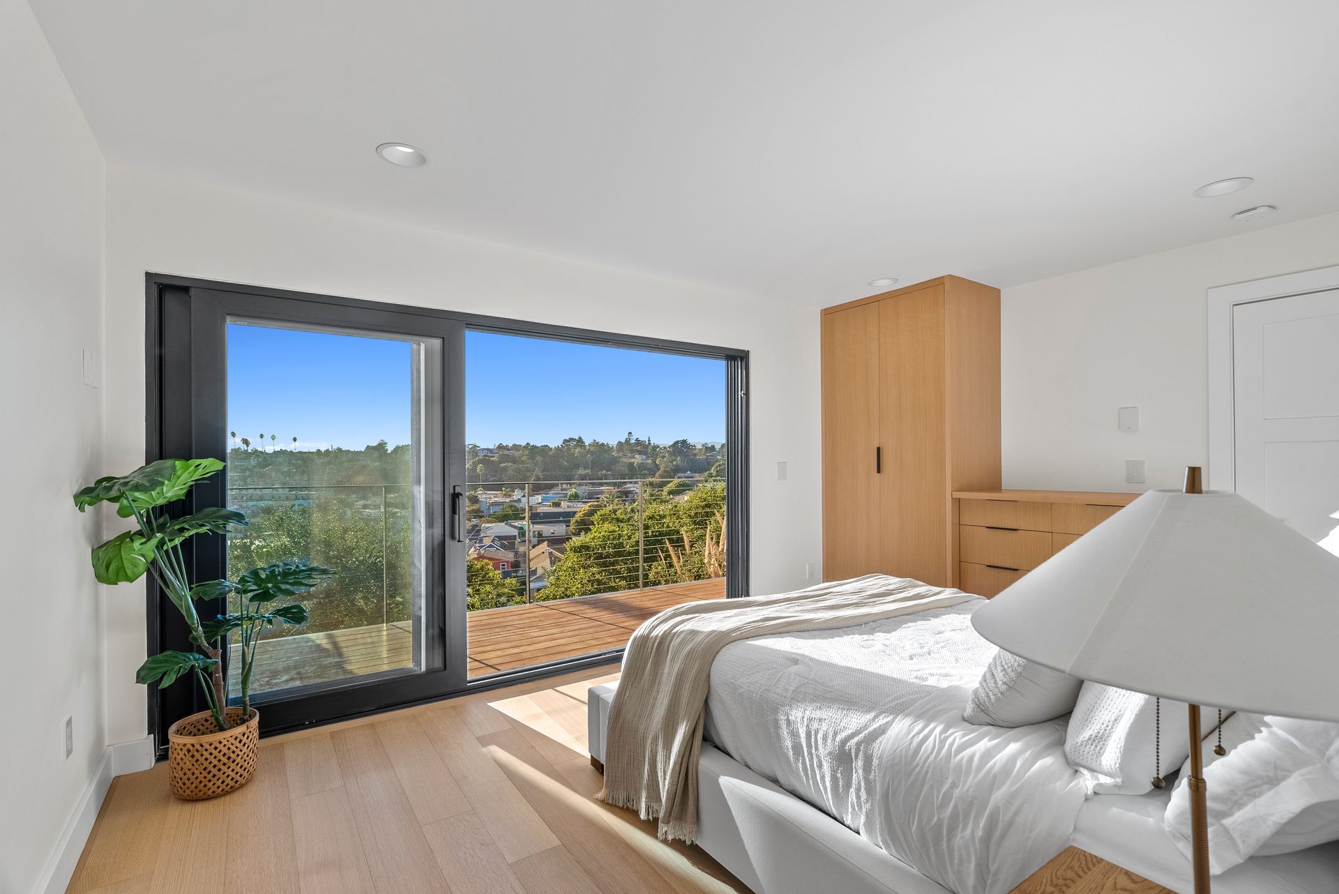Bright bedroom with a bed, potted plant, wood furniture, and sliding glass doors opening to a scenic view.