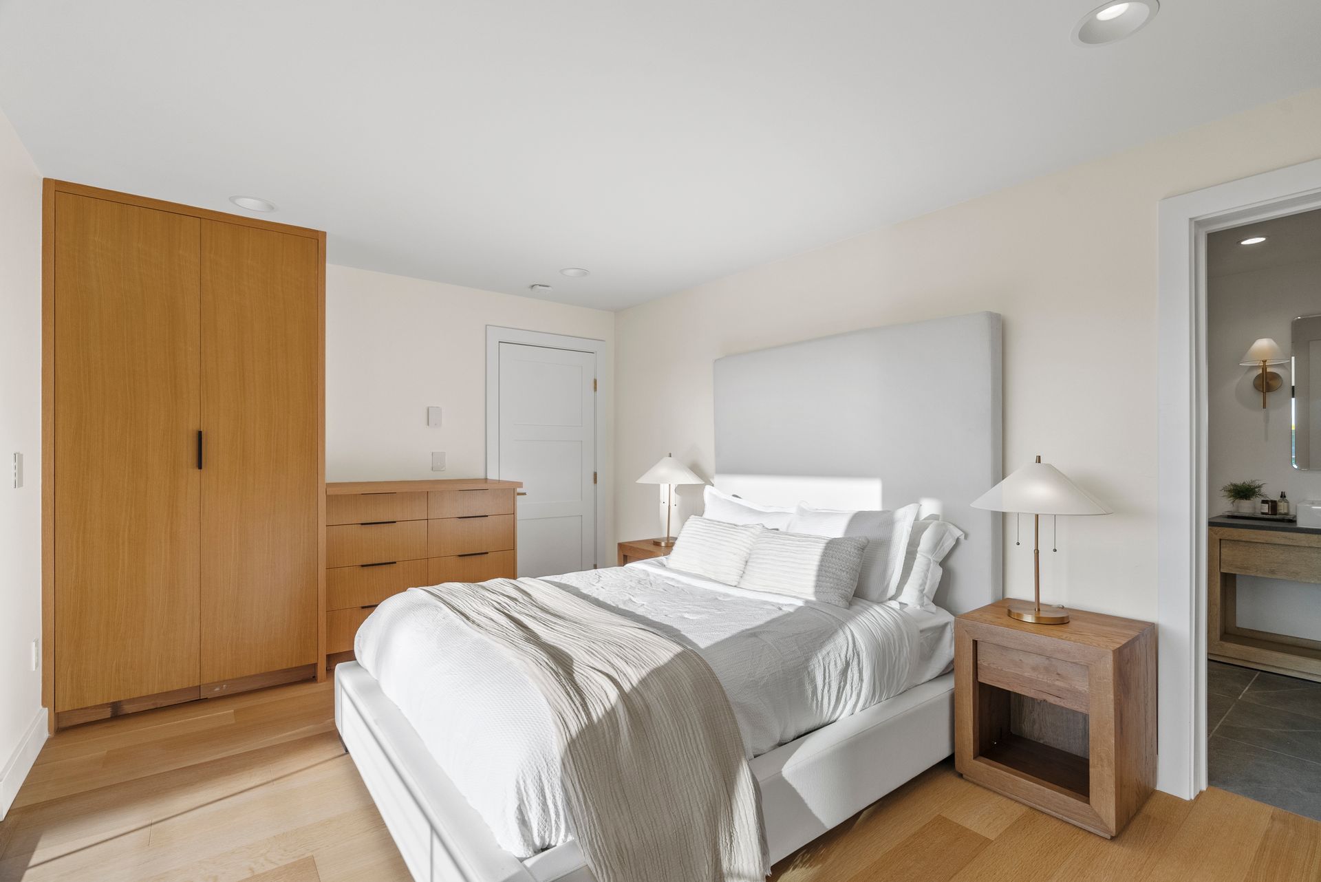 Bright bedroom with a neatly made bed, wooden dresser and wardrobe, white walls, and a bedside table with lamp.
