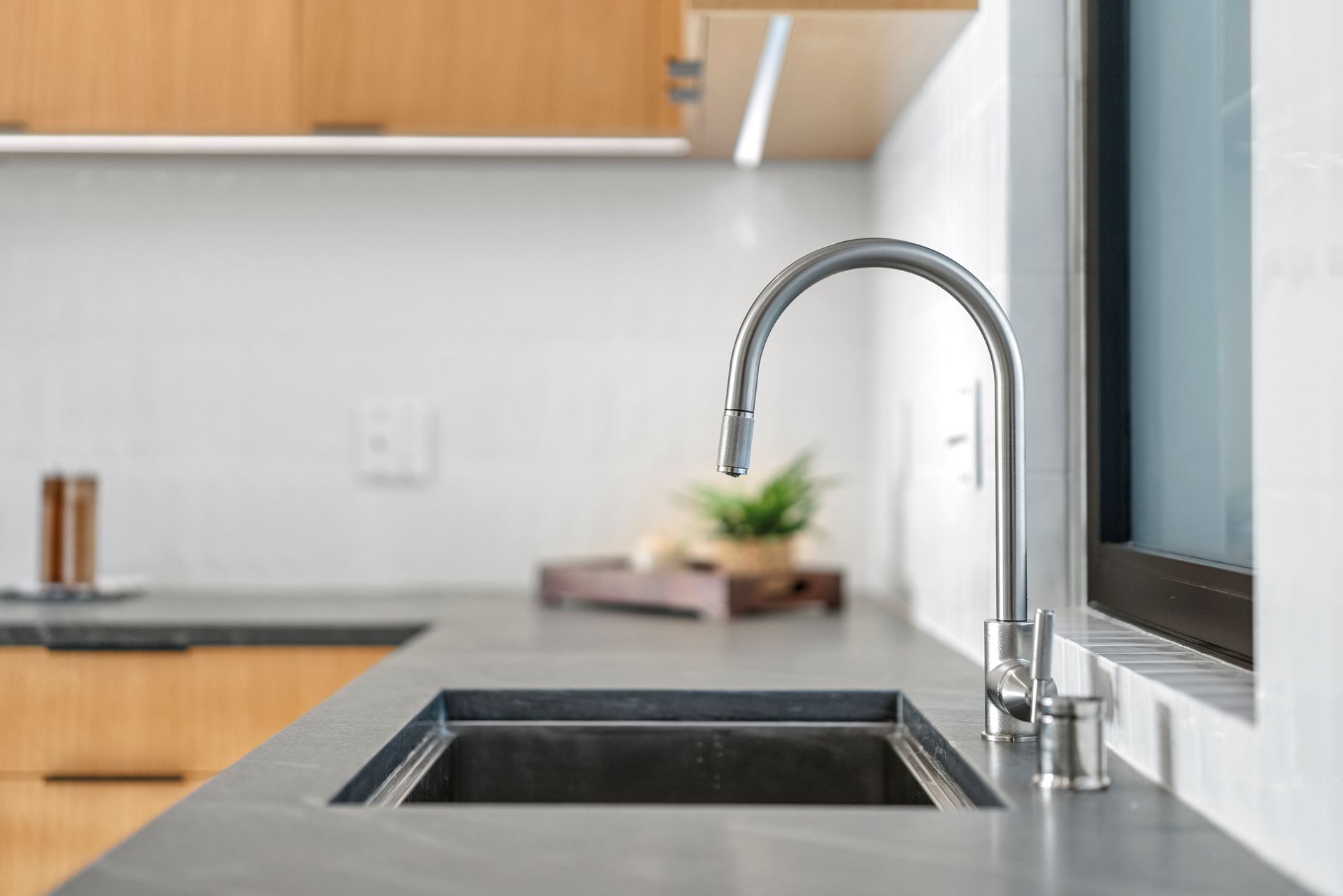 Modern kitchen sink with chrome faucet beside a window and wooden cabinets