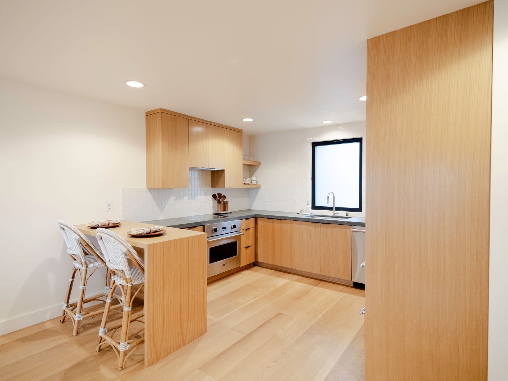 Modern kitchen with light wood cabinetry, island seating, and large window in a bright open room
