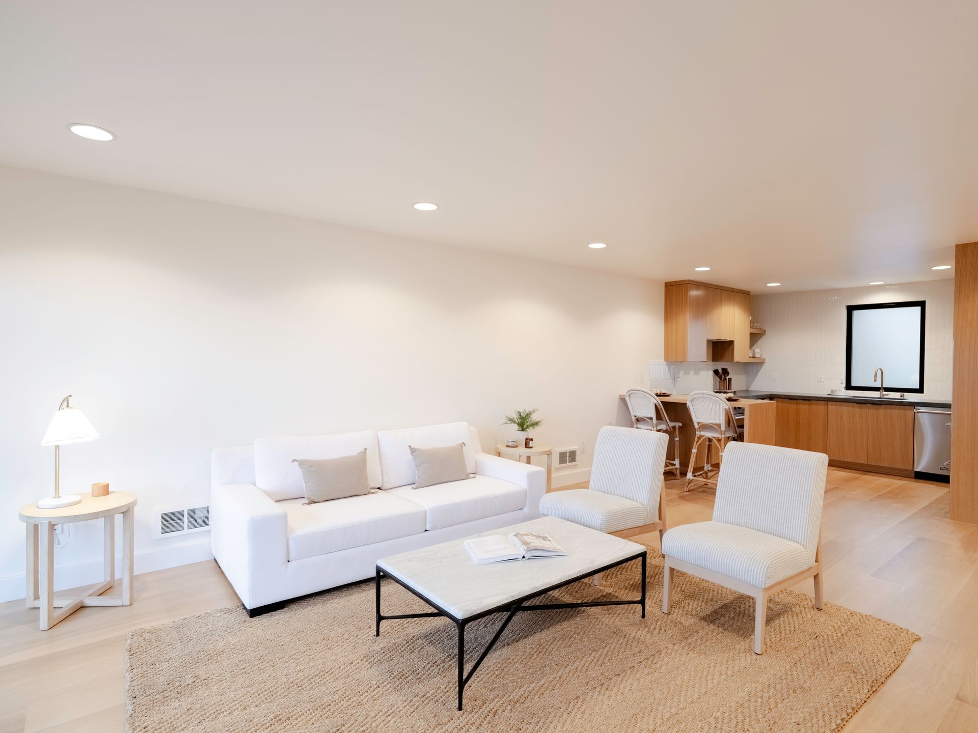 Bright modern living room with white sofa, two chairs, coffee table, and open kitchen in the background