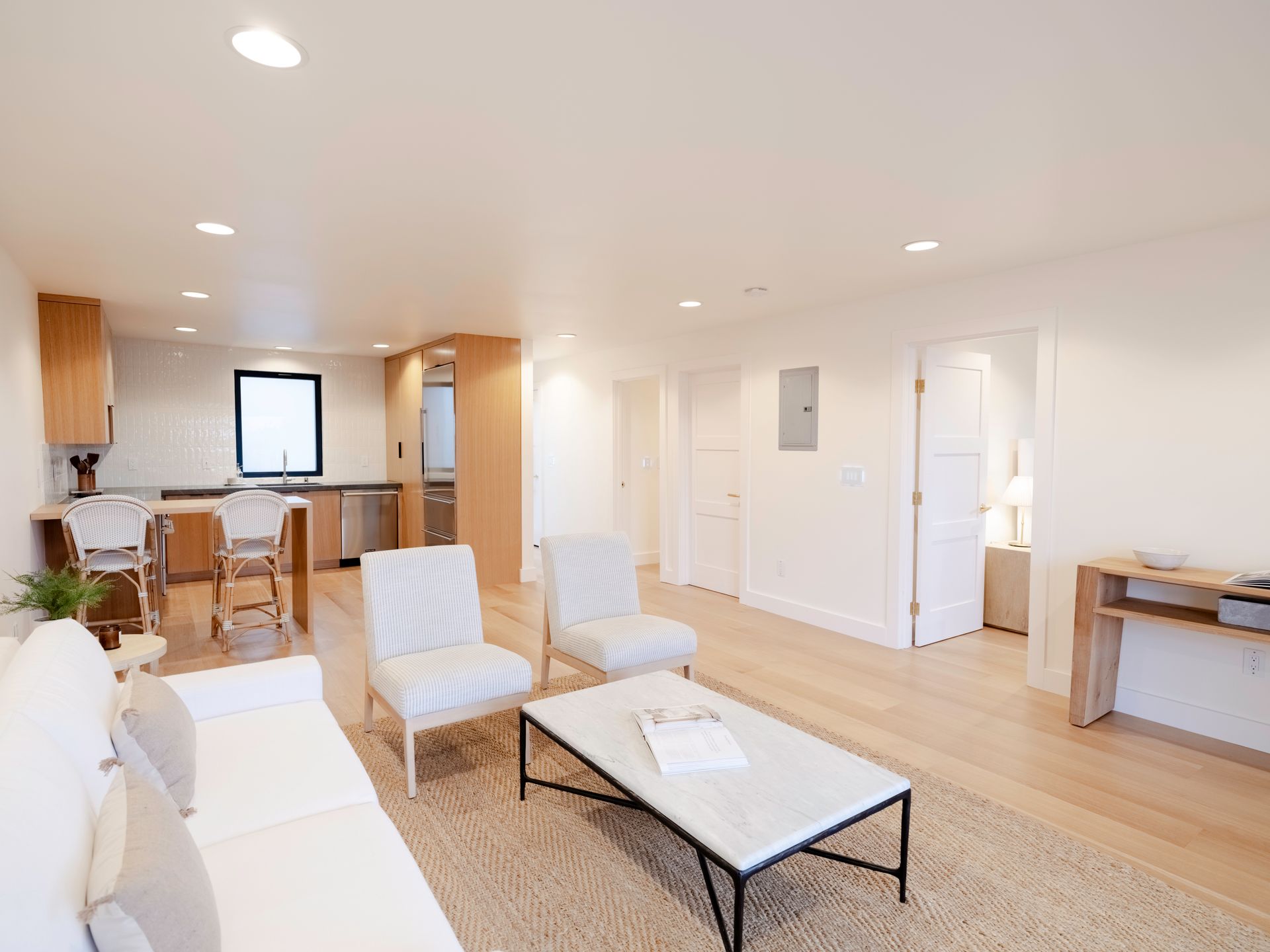 Bright open-plan living room with white furniture, wood floors, and a kitchen area in the background