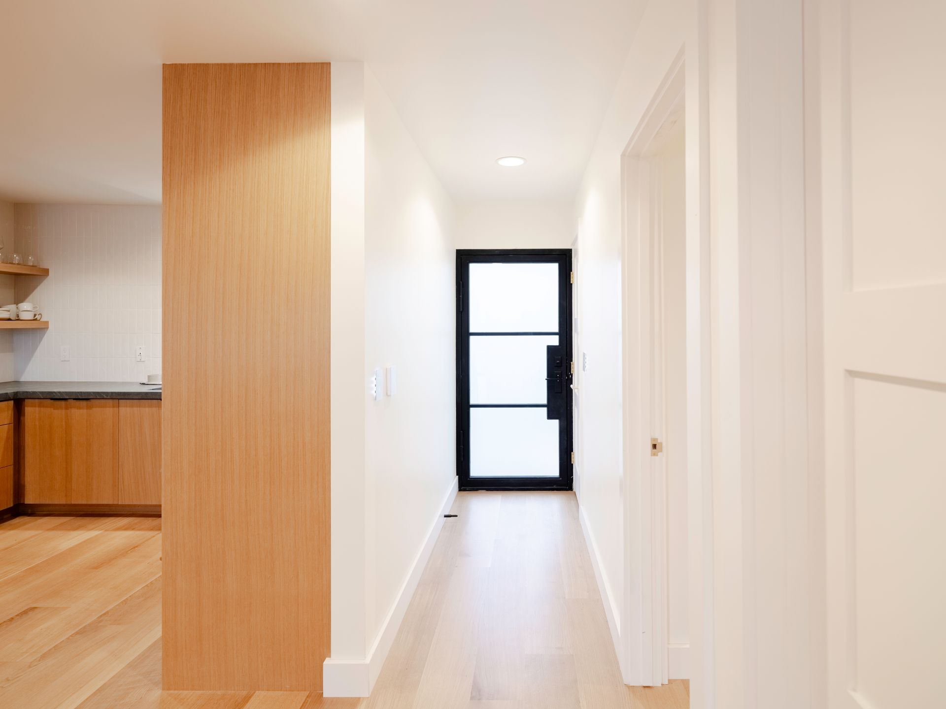Bright hallway with wood floors, white walls, and a black-framed door at the end, opening to a kitchen on the left