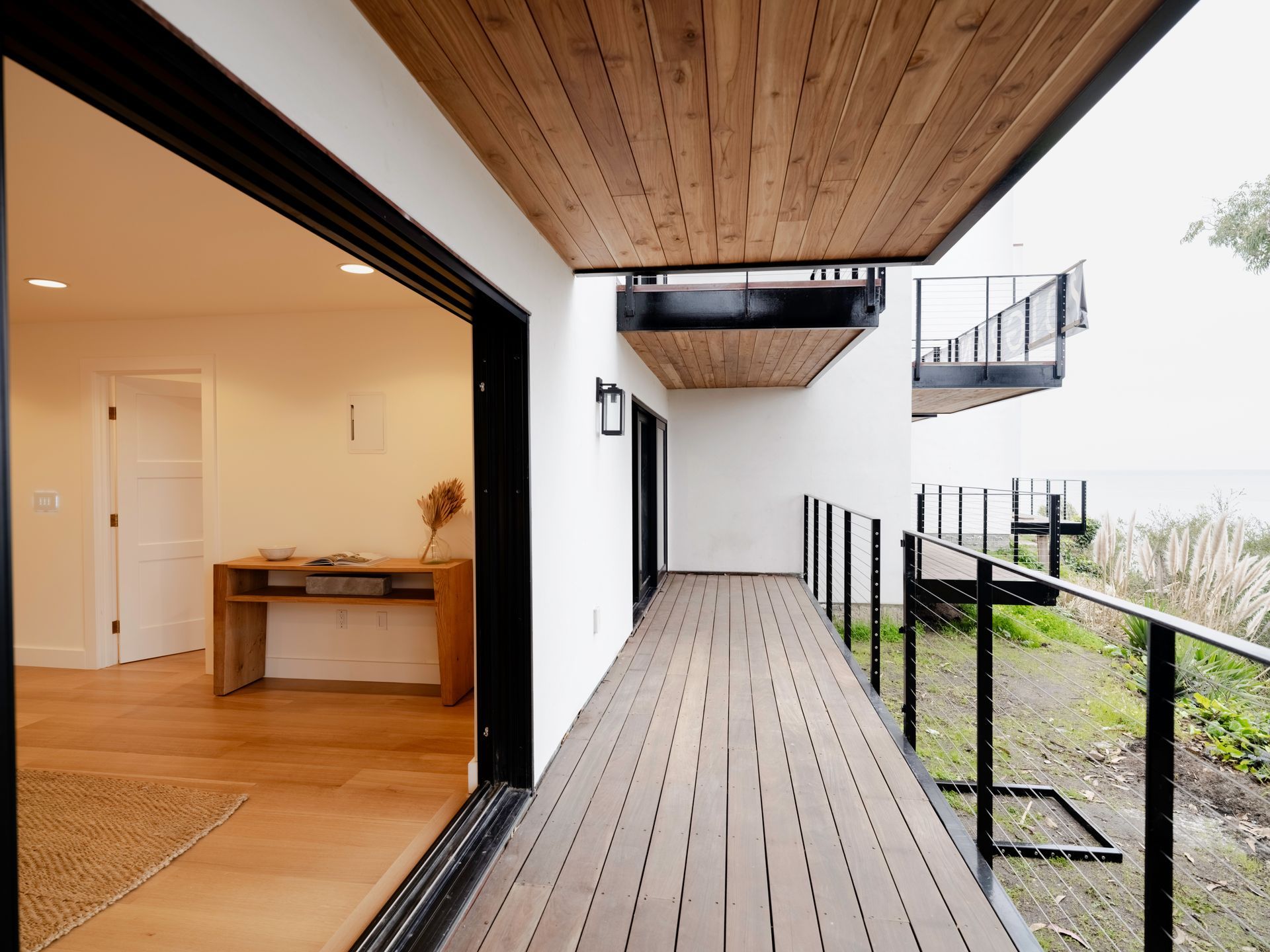 Modern wooden balcony walkway beside an open indoor room, with black railings and white exterior walls