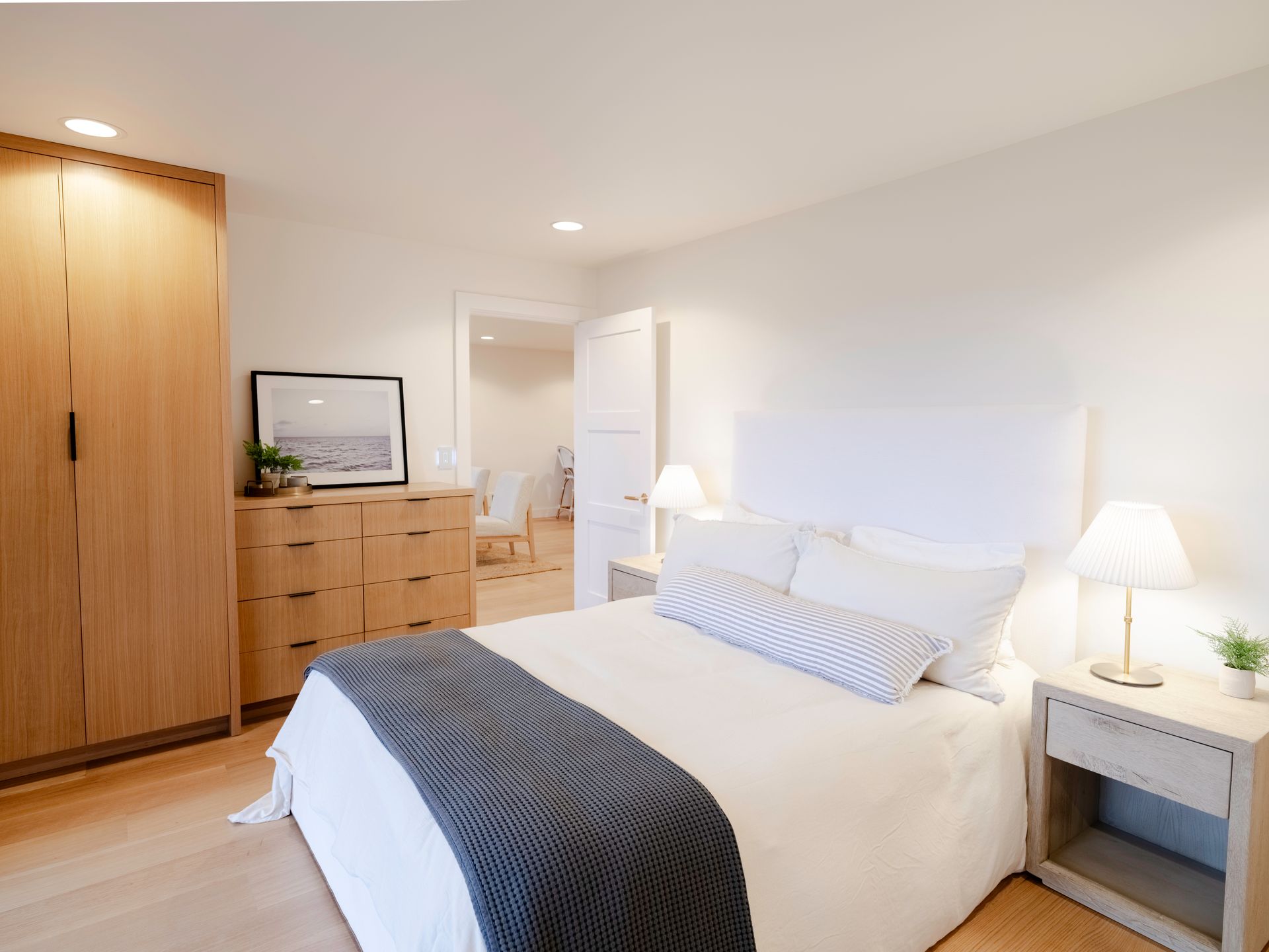 Bright modern bedroom with white bed, gray throw, bedside lamp, and wooden dresser.