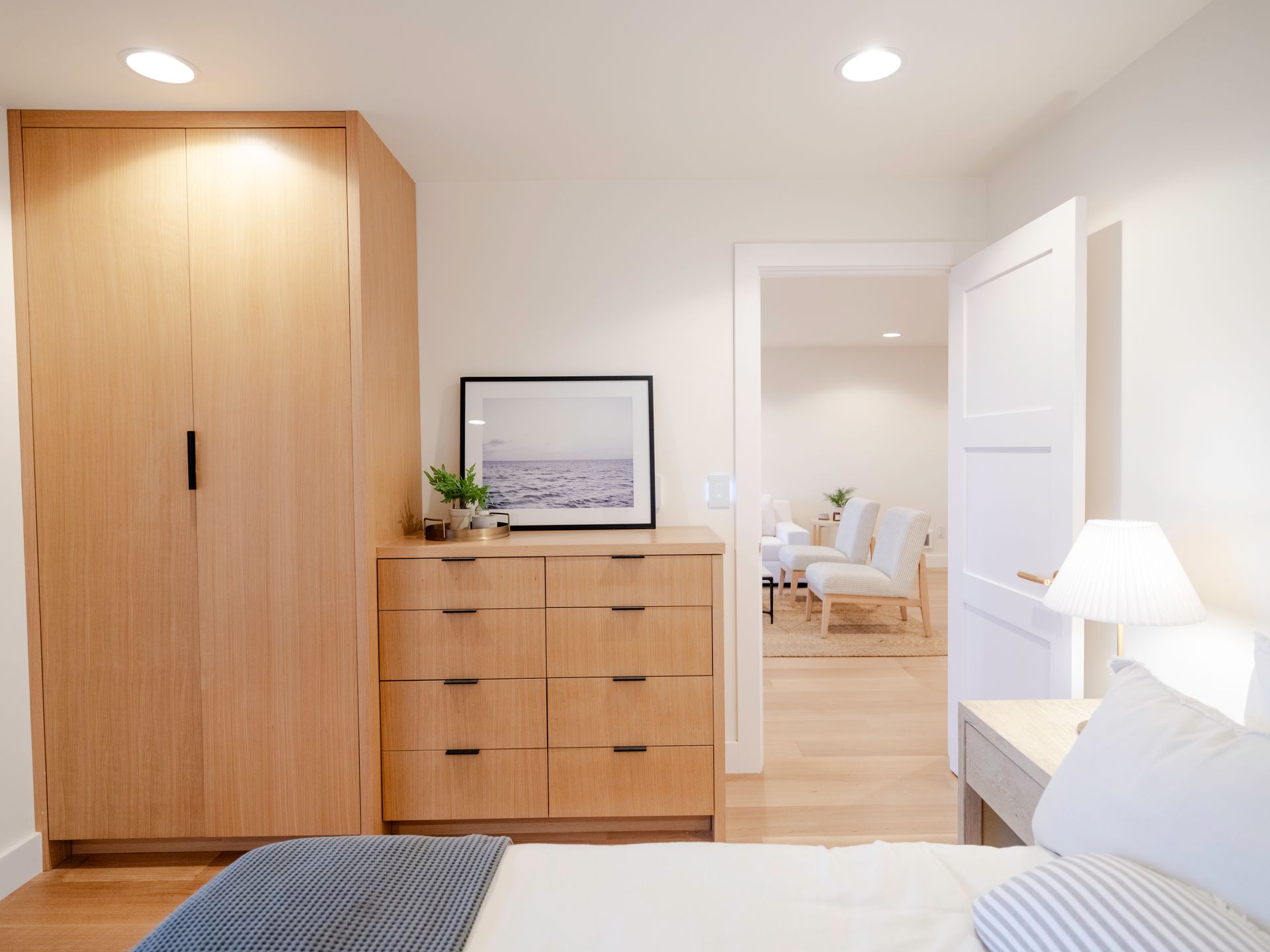 Bright bedroom with wooden wardrobe, dresser, open door to dining area, and bed corner in foreground