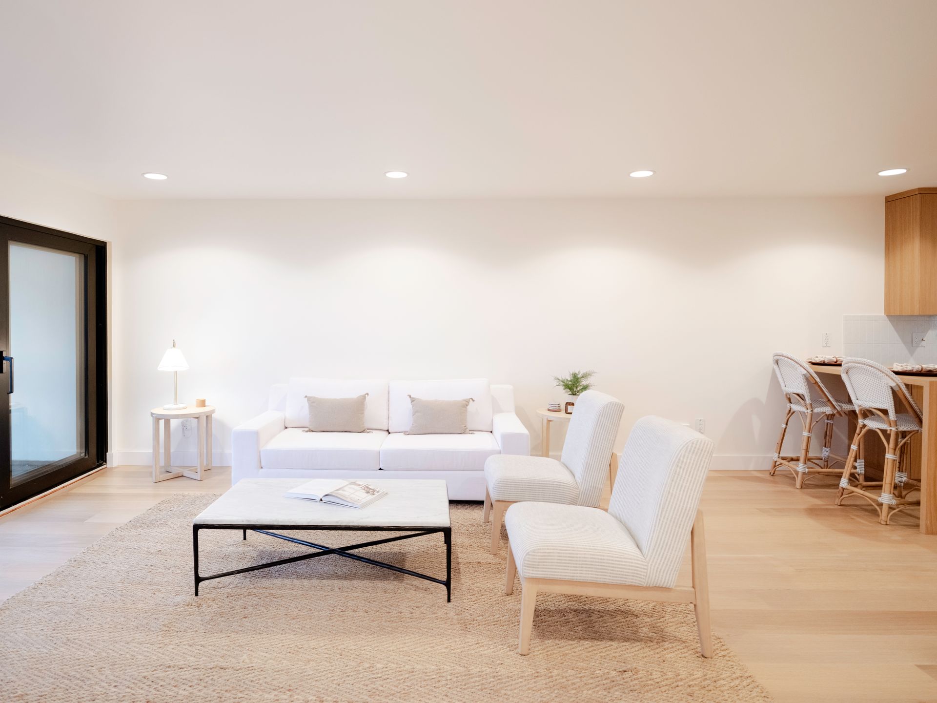 Bright minimalist living room with white sofa, chairs, coffee table, and dining area with wood flooring.