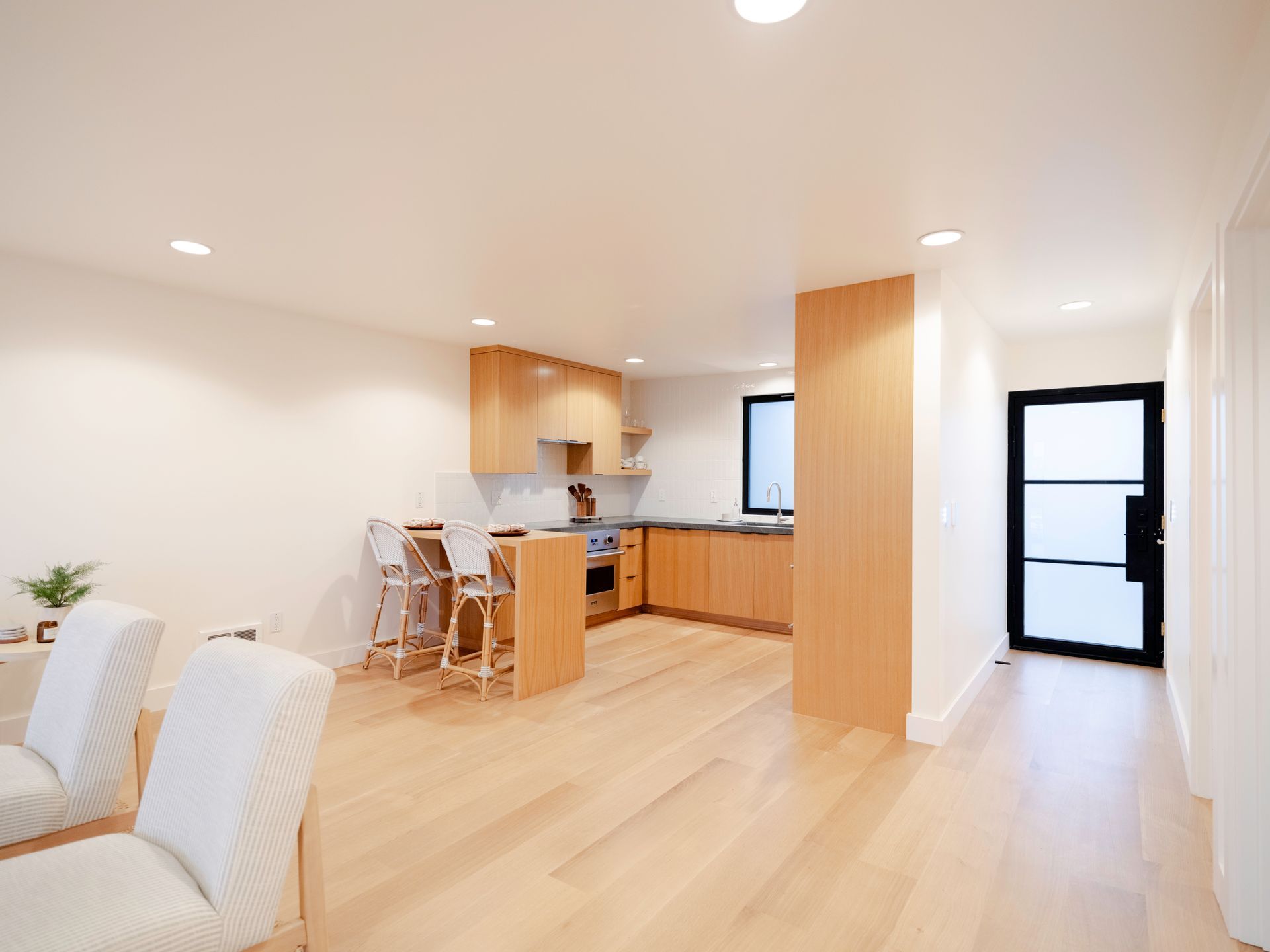 Bright open-plan living room and kitchen with light wood floors, white walls, and a black-framed door.