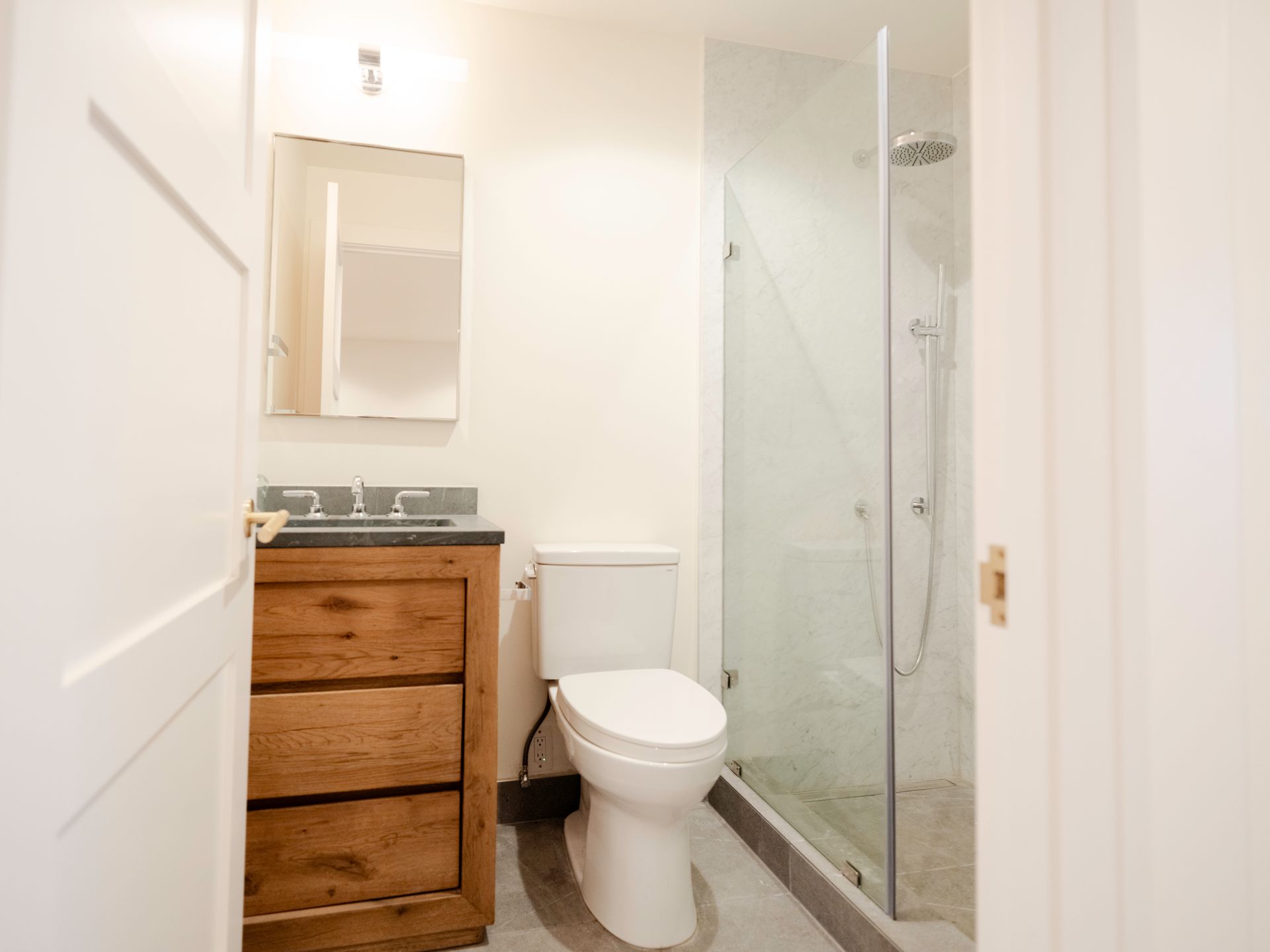 Small bathroom with wooden vanity, toilet, and glass-enclosed shower beside a mirror.