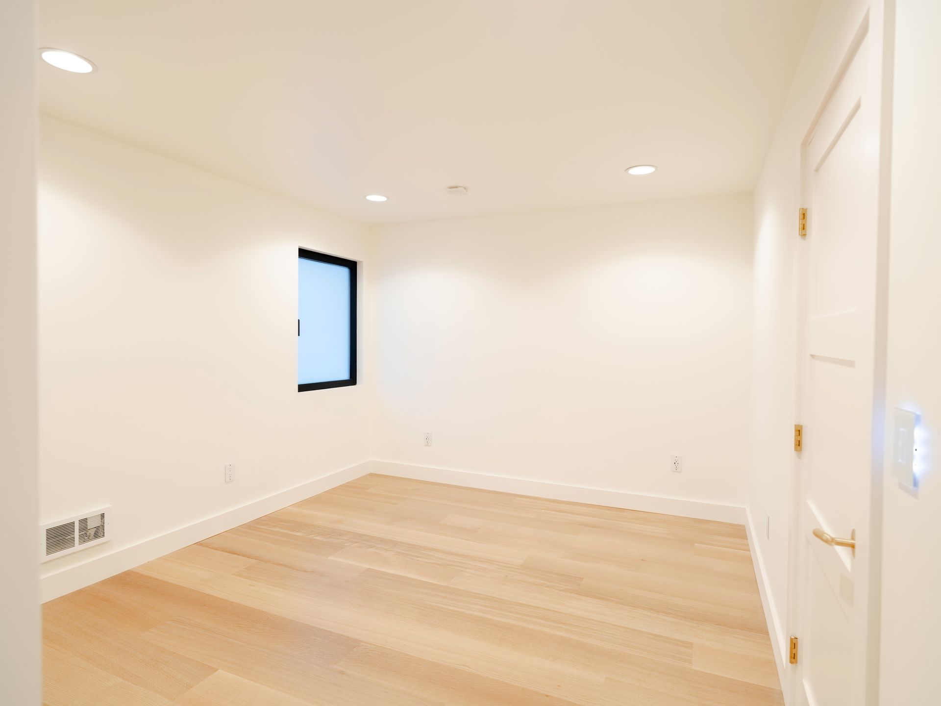 Empty white room with light wood floors, recessed lights, and a small window.