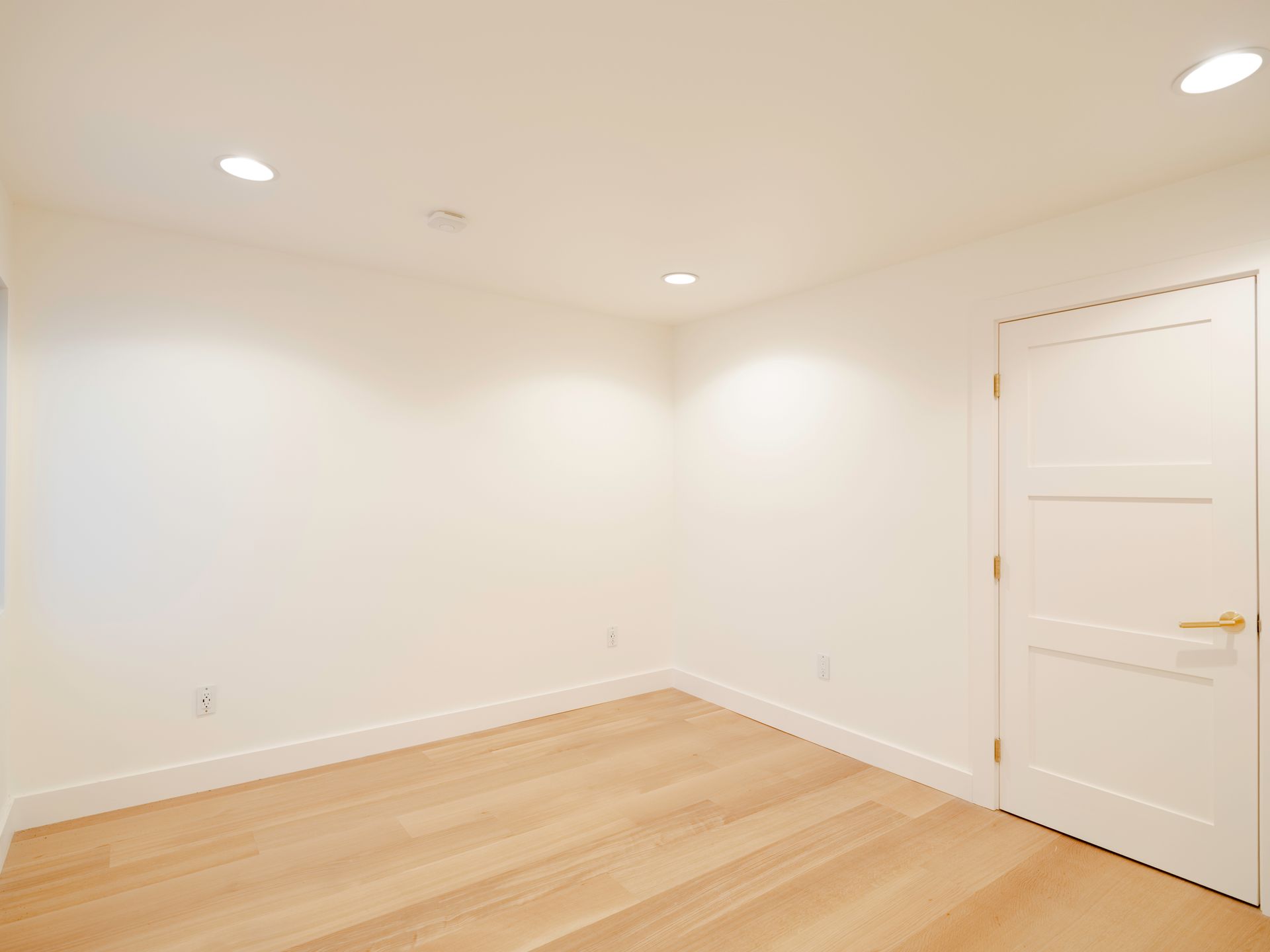 Empty white room with light wood floor, recessed ceiling lights, and a closed white door